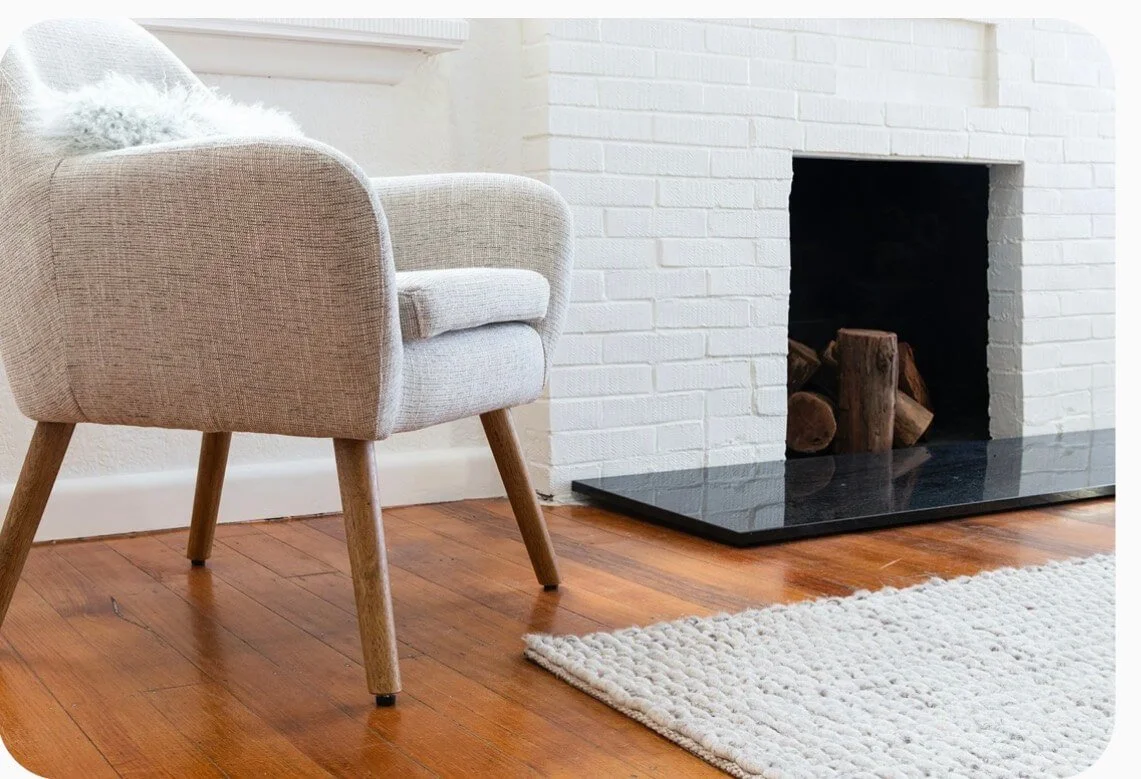 A cozy living room corner with a beige upholstered armchair, a white fluffy pillow, a white textured rug, and a modern black fireplace with logs inside, set on a wooden floor.