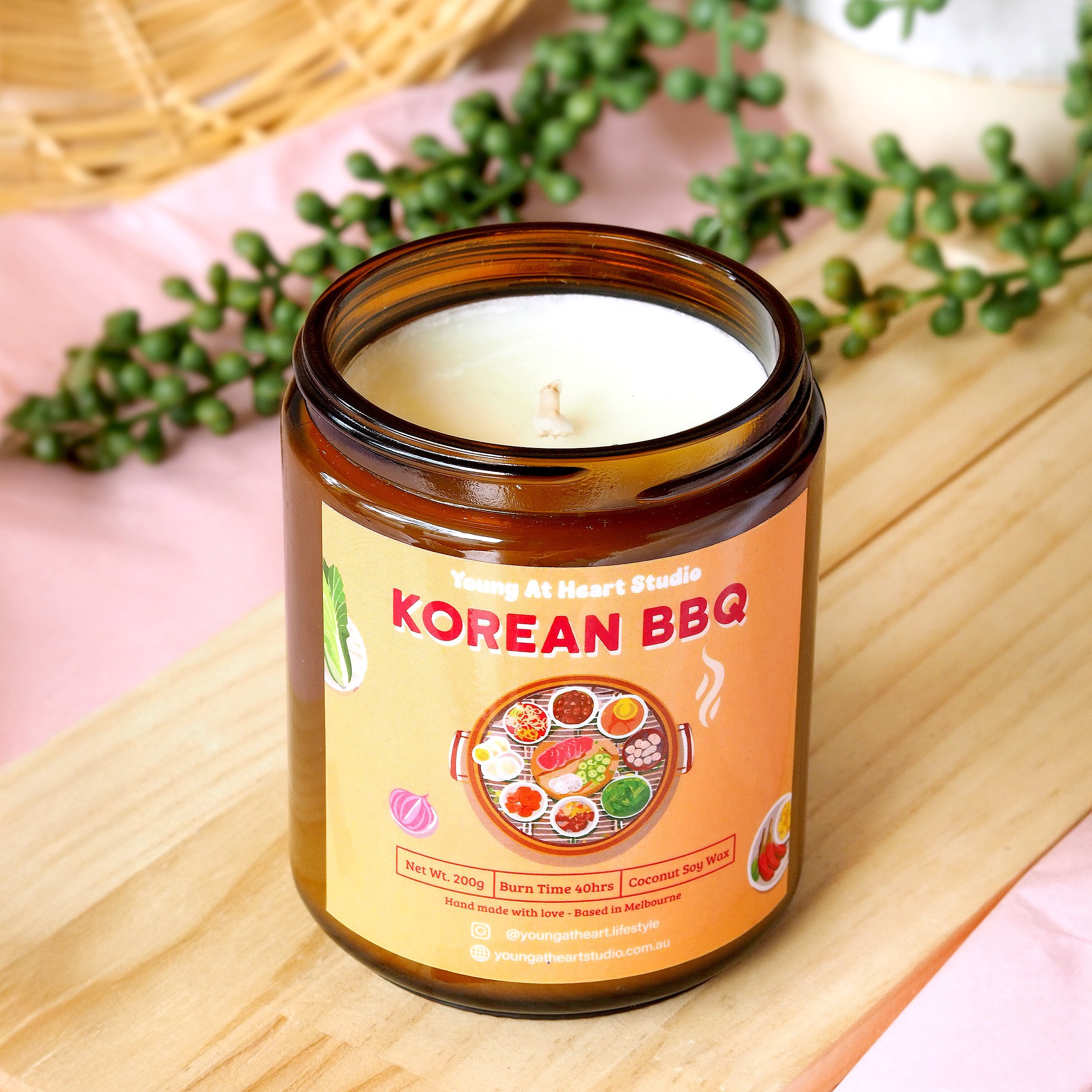 A red container with a gold lid labeled "Korean BBQ" and the words "Young At Heart Studio" at the top. The label features images of Korean foods, a candle fact section, and a description of the product.