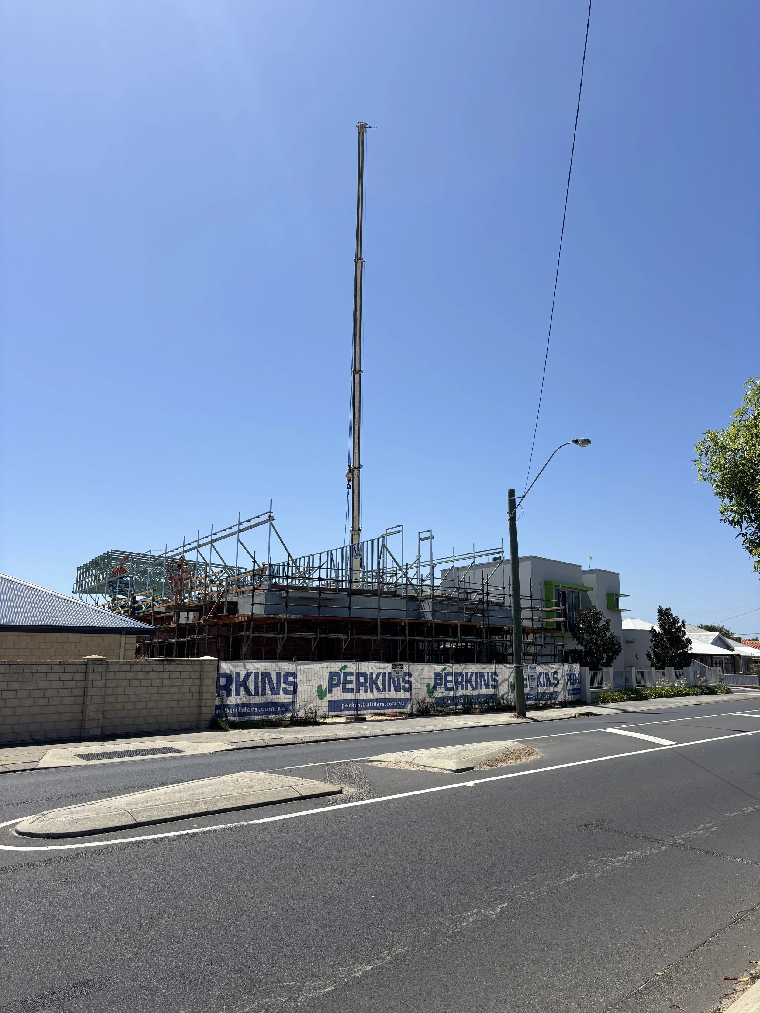 Construction site of a building with scaffolding, a crane, and a clear blue sky. The site is surrounded by a brick wall with banners reading Perkins. Street view with a sidewalk and a streetlight, some trees, and part of a building with a green and white exterior.