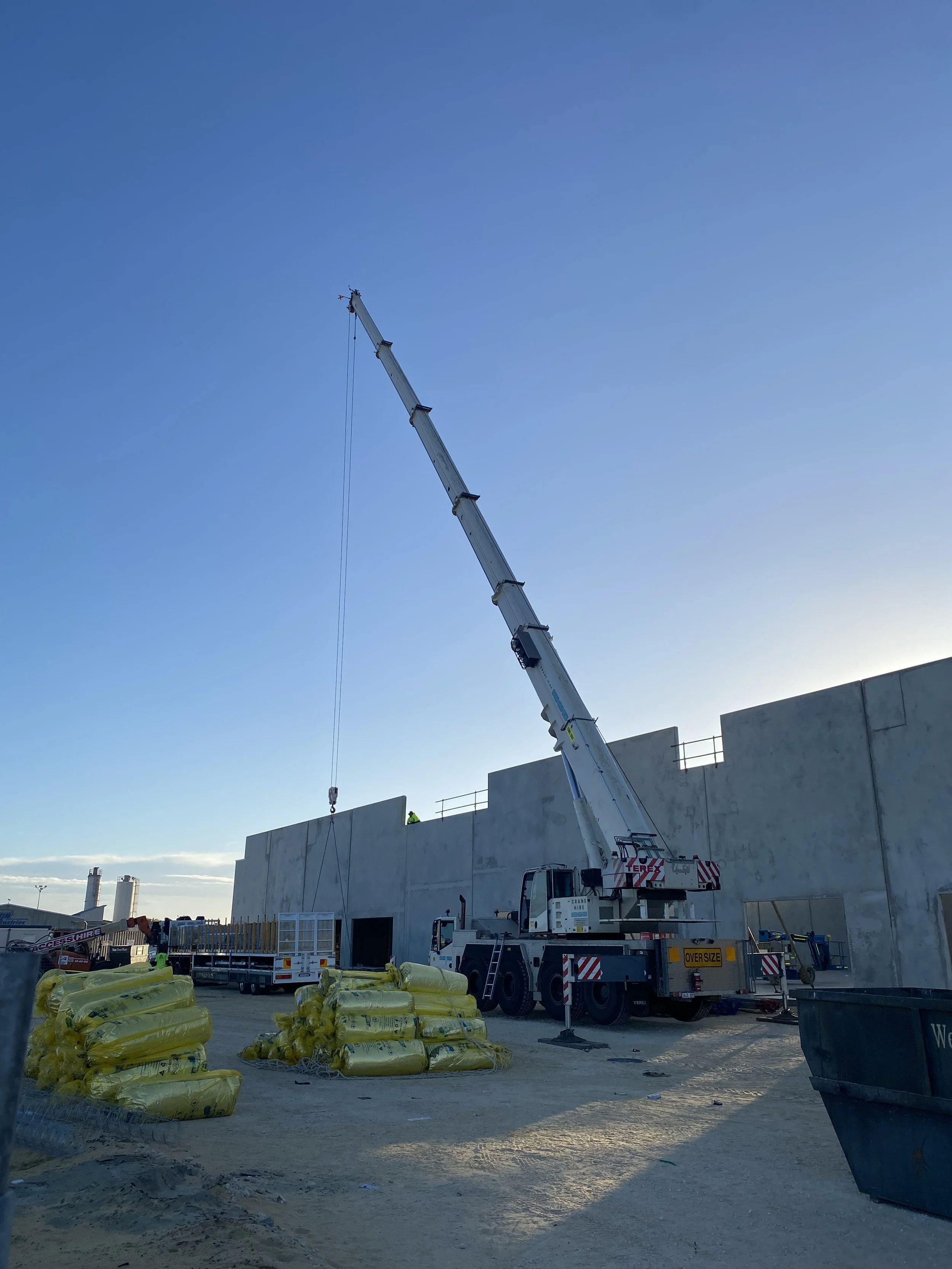 Construction site with a large white crane extending upward, workers in safety gear, yellow wrapped construction materials on the ground, and a large gray concrete building under construction.