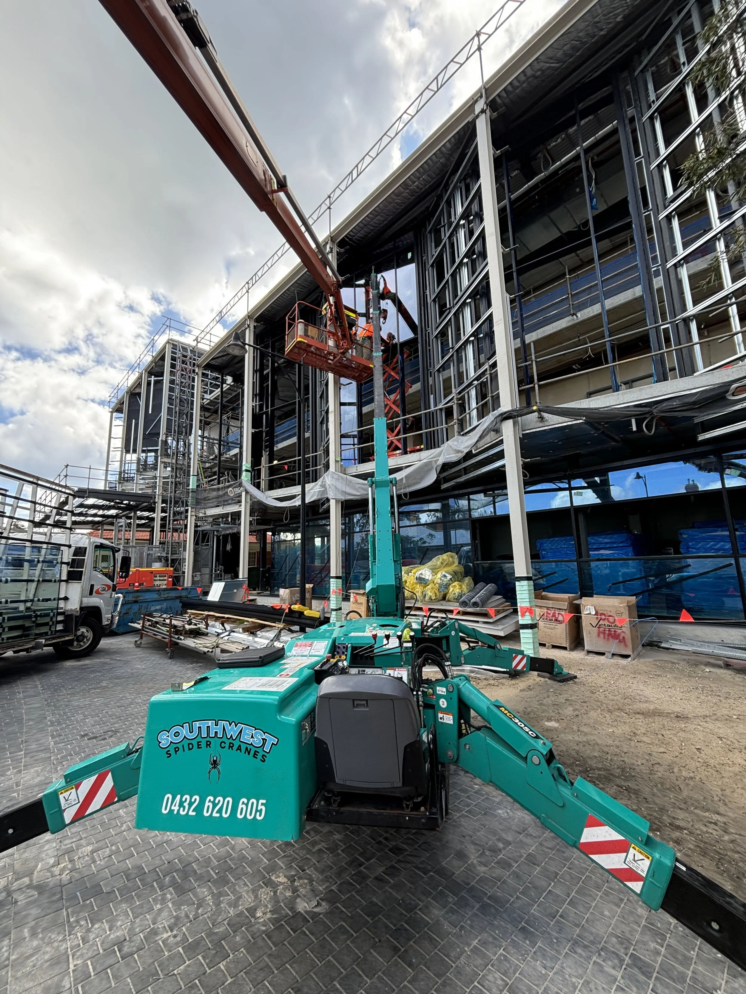 Construction site with a worker on a lift installing or repairing a building's exterior. The building has scaffolding and workers inside. Construction equipment, materials, and a small truck are in the foreground.