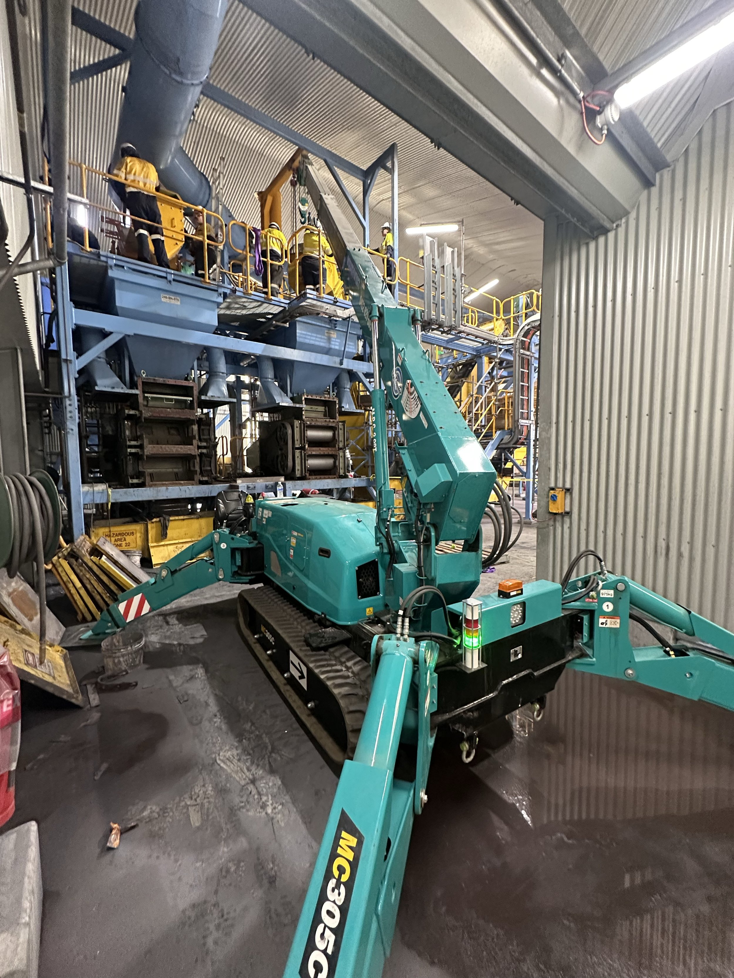 Inside an industrial facility, a teal compact excavator with tracks and an extended arm is positioned near a metal wall, surrounded by machinery, hoses, and tools, with workers in yellow safety gear on a raised platform working on equipment overhead.