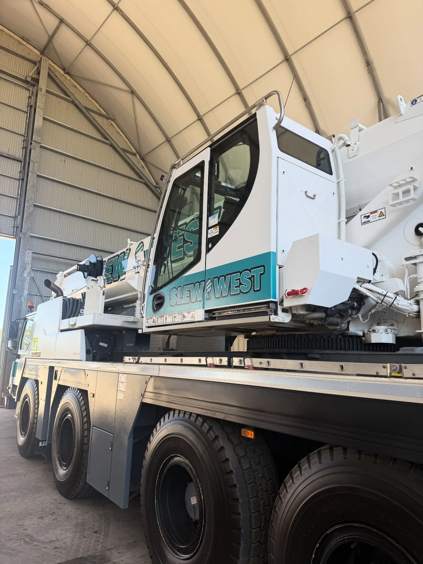A large white crane with the brand name Slew West is mounted on a black truck trailer inside a warehouse with a curved metal roof.