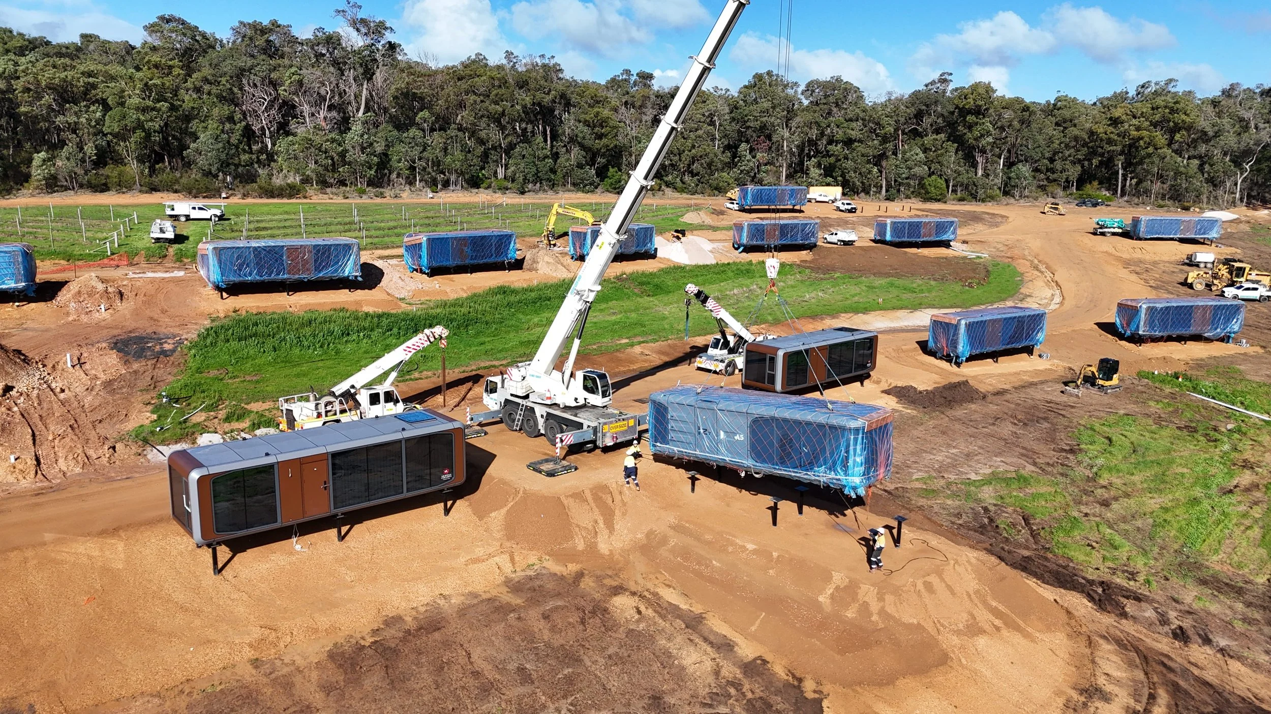 Construction site with multiple large mobile units covered in blue protective wrapping, heavy machinery including cranes and excavators, and trucks on a dirt road, with forest in the background.