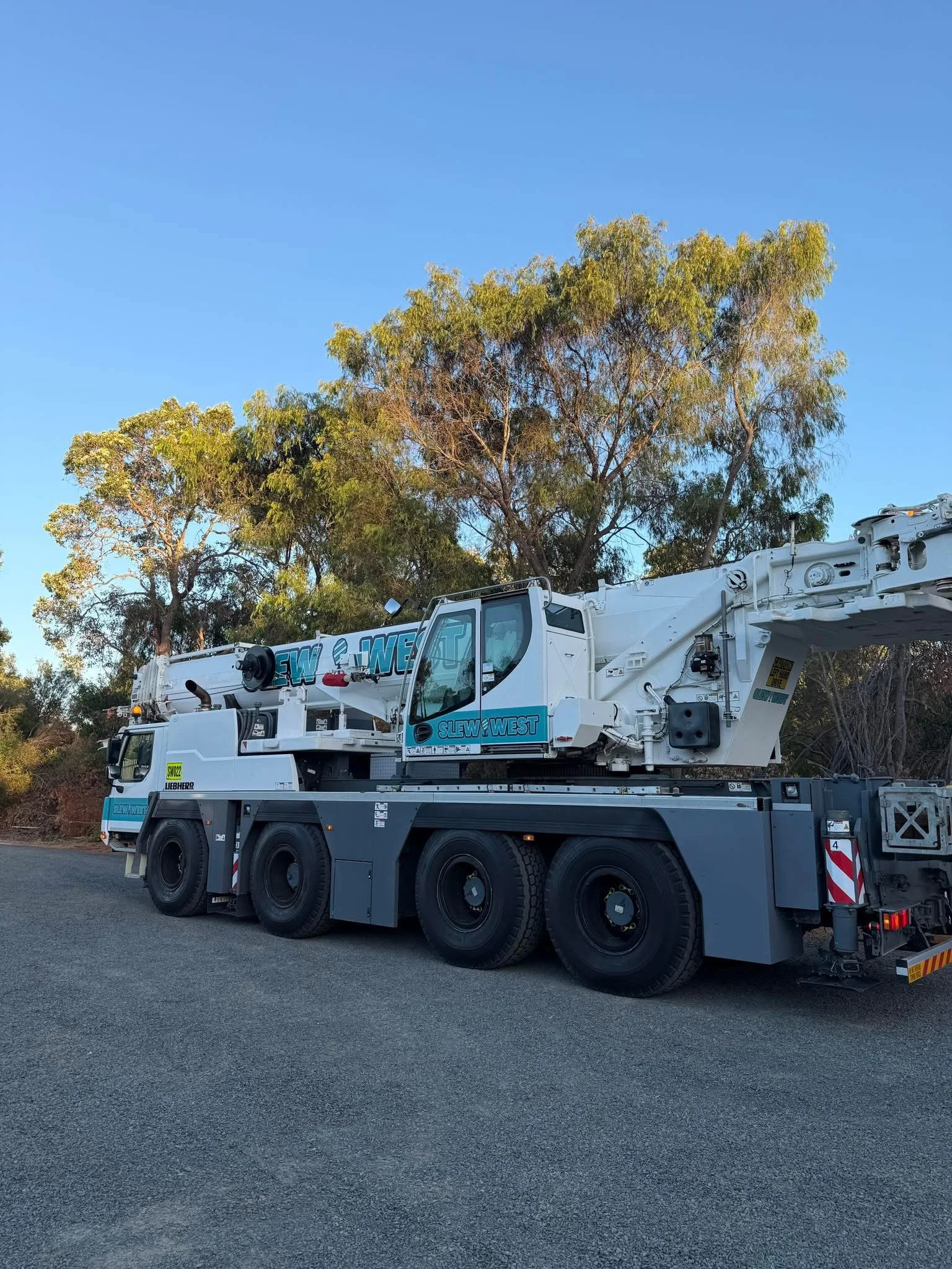 A white mobile crane truck parked on a paved road with trees and a blue sky in the background.
