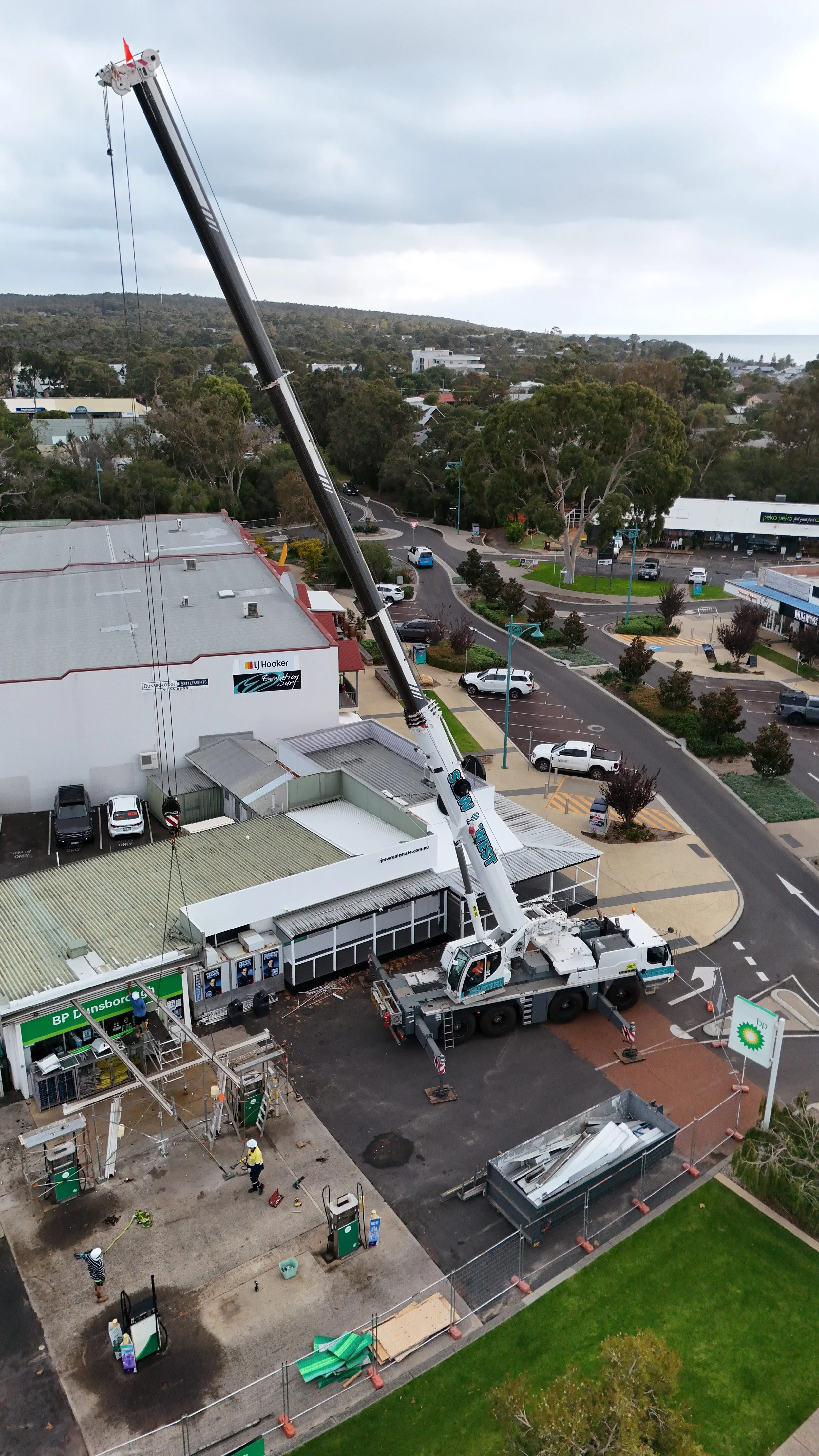 A construction crane is lifting materials or equipment to the top of a building. Construction workers are working on the ground, and nearby are parked cars, commercial establishments, and a small green park area.