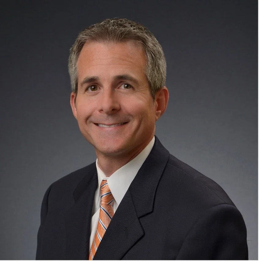 Professional portrait of a middle-aged man in a suit with a white shirt and orange striped tie, smiling against a dark background.