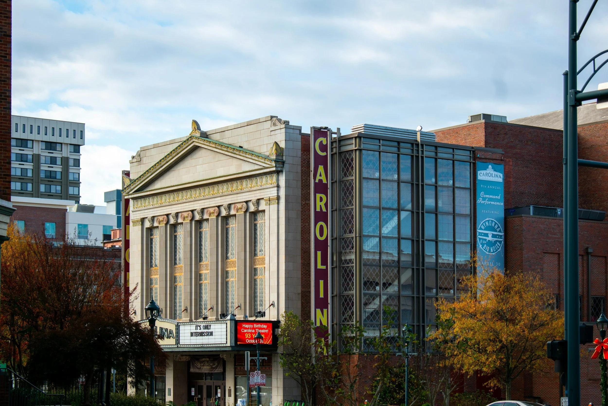 The Carolina Theatre building with a marquee and tall signs, adjacent to trees with autumn-colored leaves, in an urban setting with other buildings in the background.