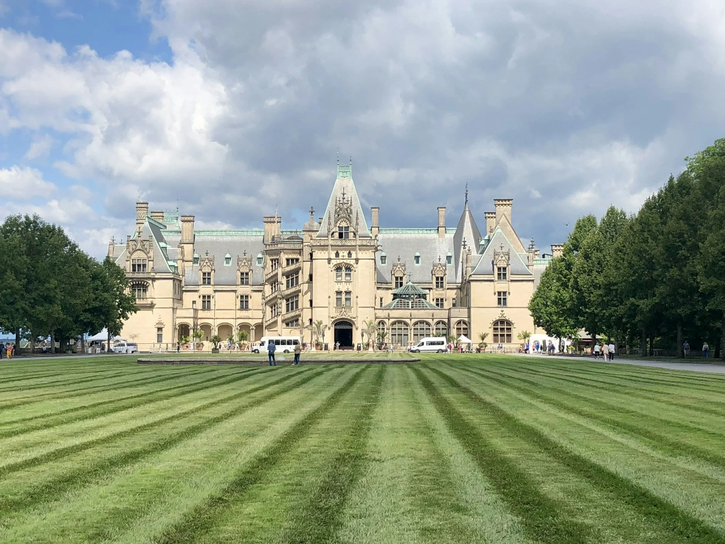 A large, ornate castle with multiple towers and turrets, surrounded by a well-manicured lawn with striped grass, trees on either side, and a cloudy sky overhead.