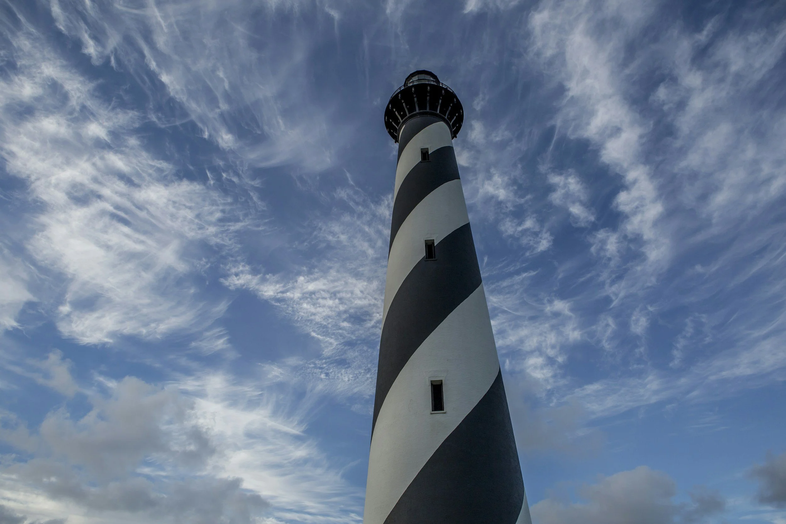 A tall lighthouse with black and white spiral stripes against a partly cloudy sky.