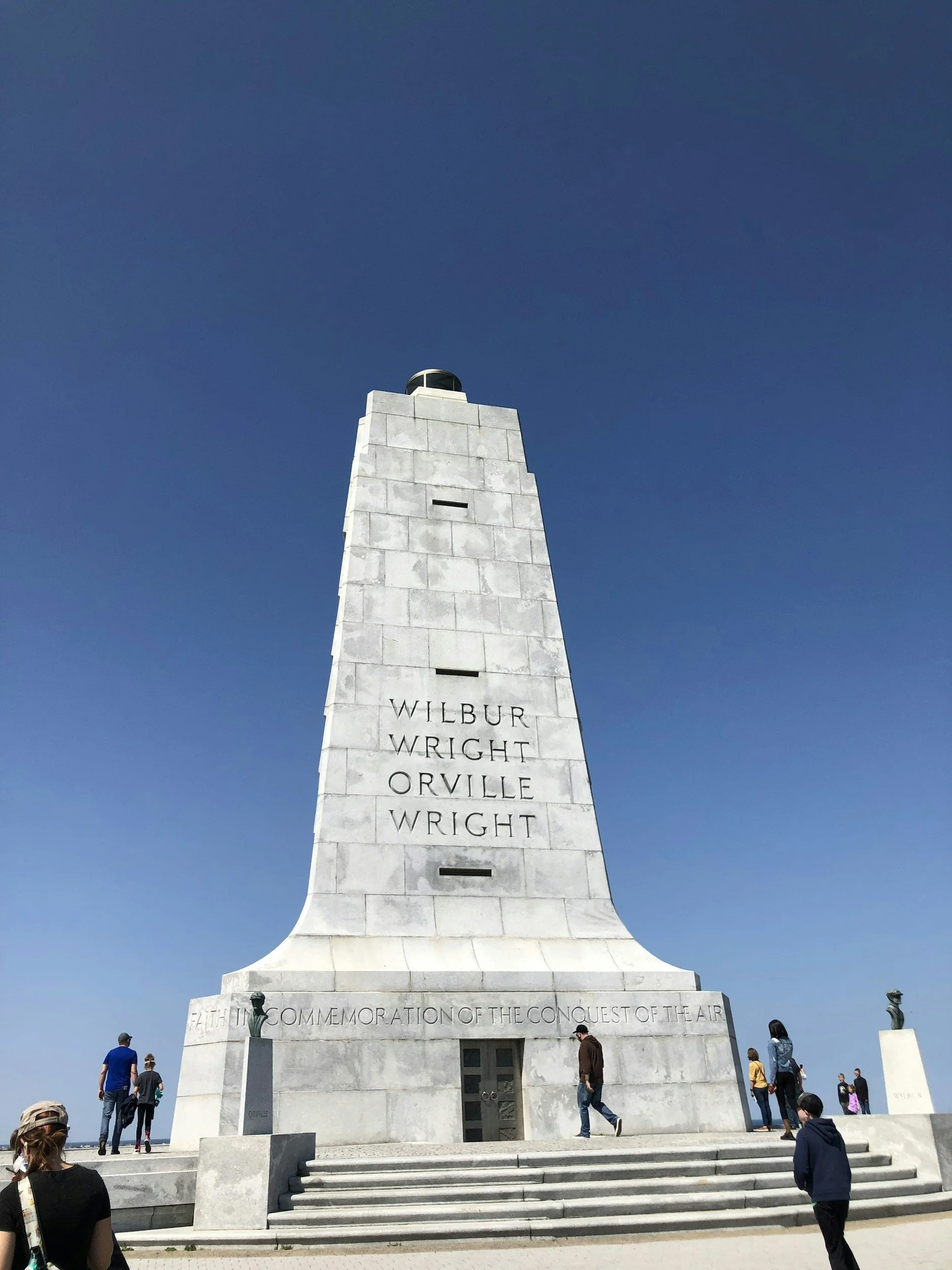 A tall white memorial monument dedicated to Wilbur Wright, with several people visiting, set against a clear blue sky.