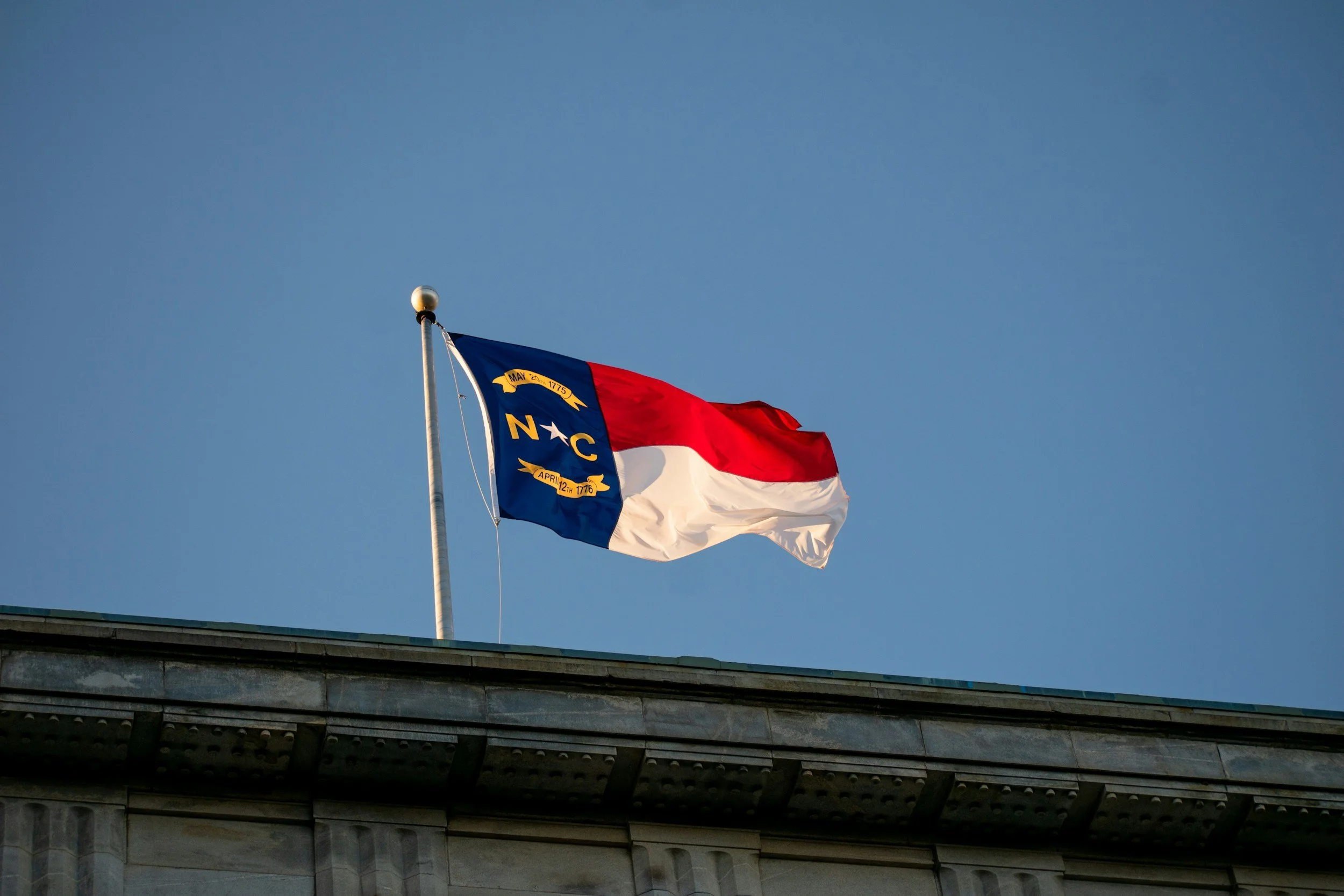 North Carolina state flag flying on a flagpole above a building against a blue sky.