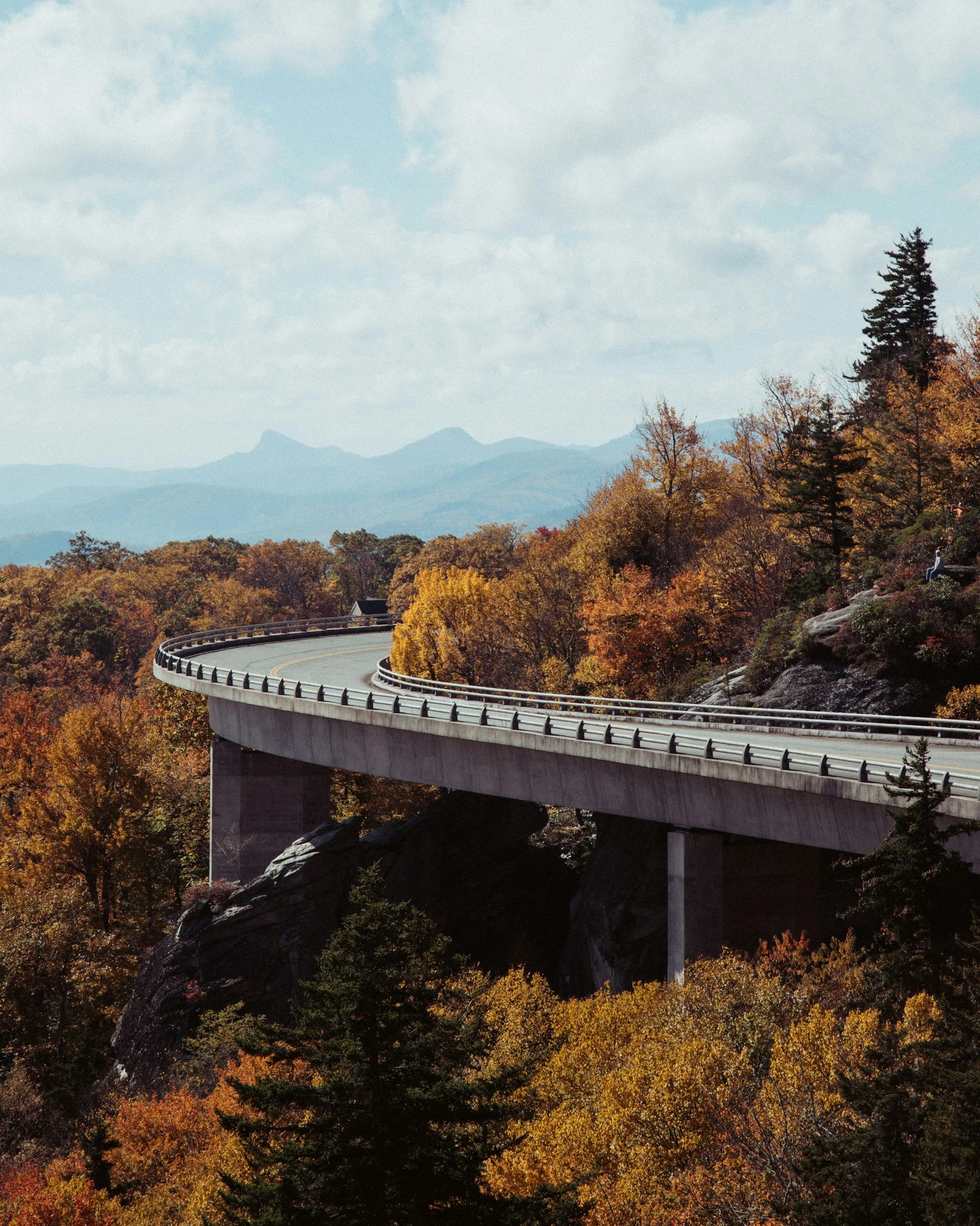 Landscape of rolling hills covered with fall foliage in various shades of orange, yellow, and red, with large rocks prominent on a hillside, in the distance layers of mountains shrouded in mist.