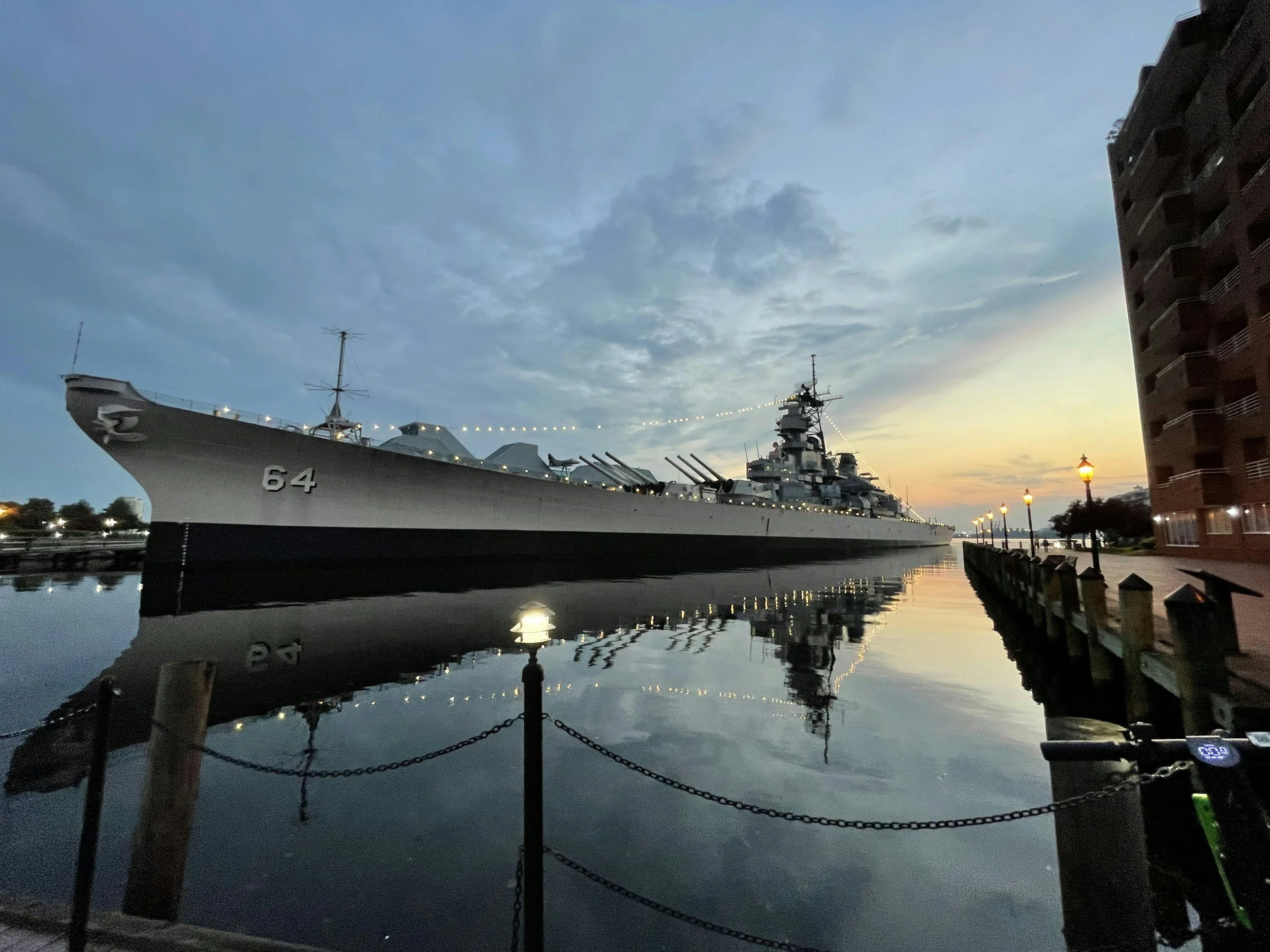 A large gray battleship with the number 64 on its side docked at a pier during dusk, with calm water reflecting the ship and a few lit lampposts along the walkway.