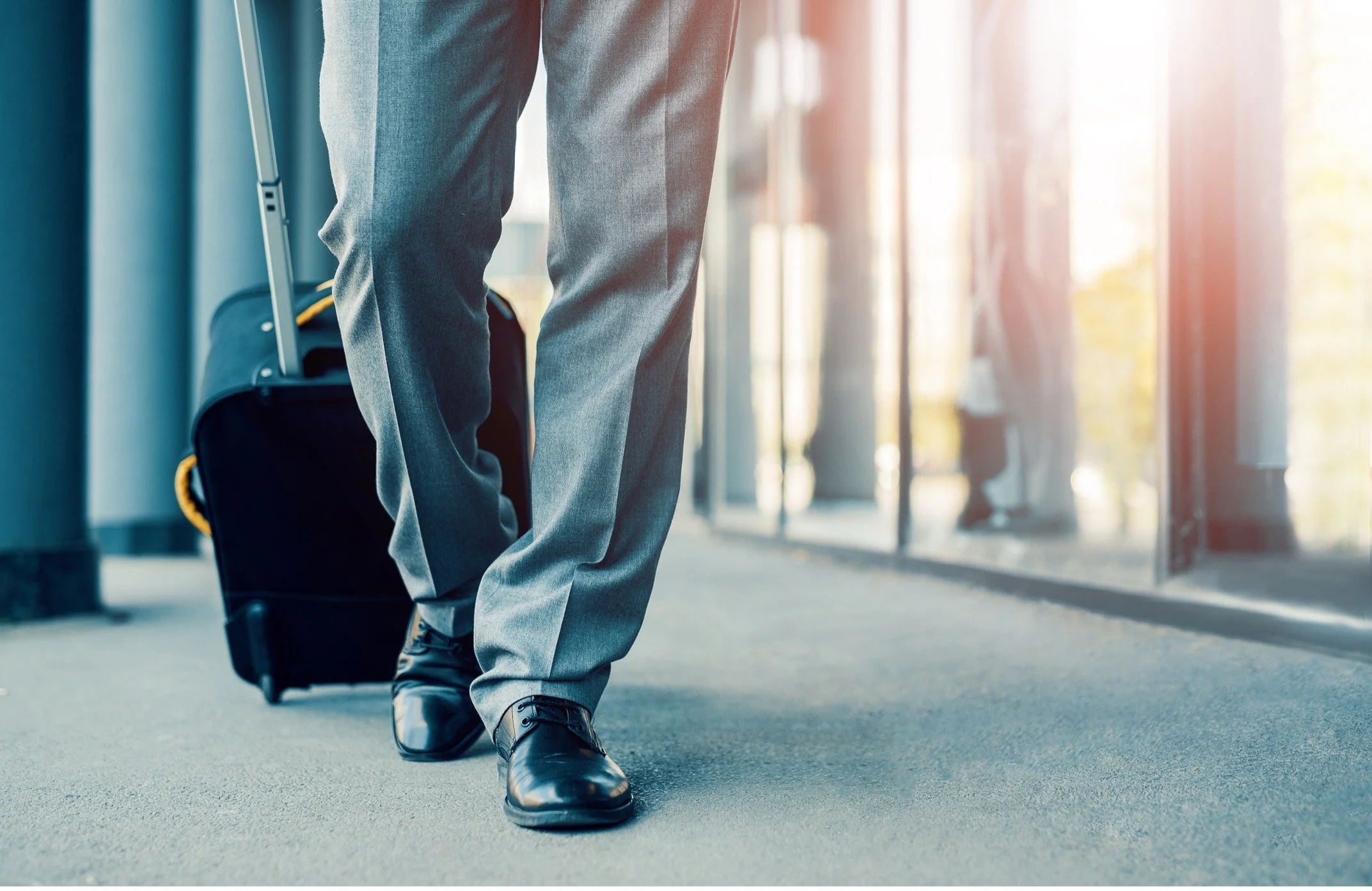 A person walking through an airport terminal with a rolling suitcase, wearing formal gray pants and black shoes, with sunlight streaming through large windows.