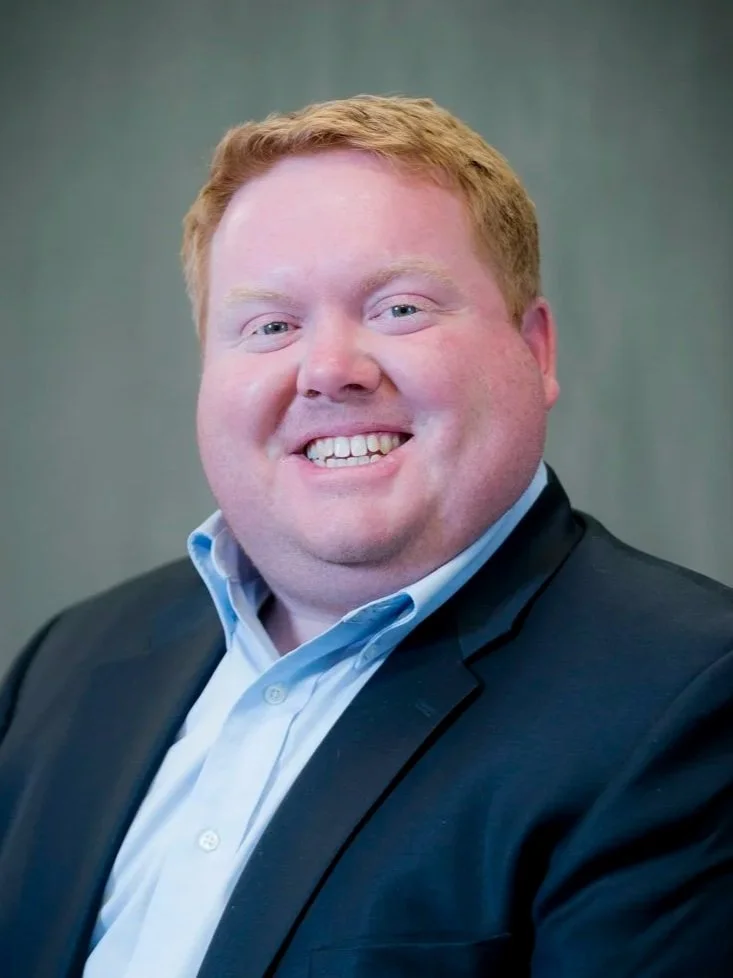 A smiling man with red hair and fair skin in a professional setting, wearing a dark blazer and light blue dress shirt.