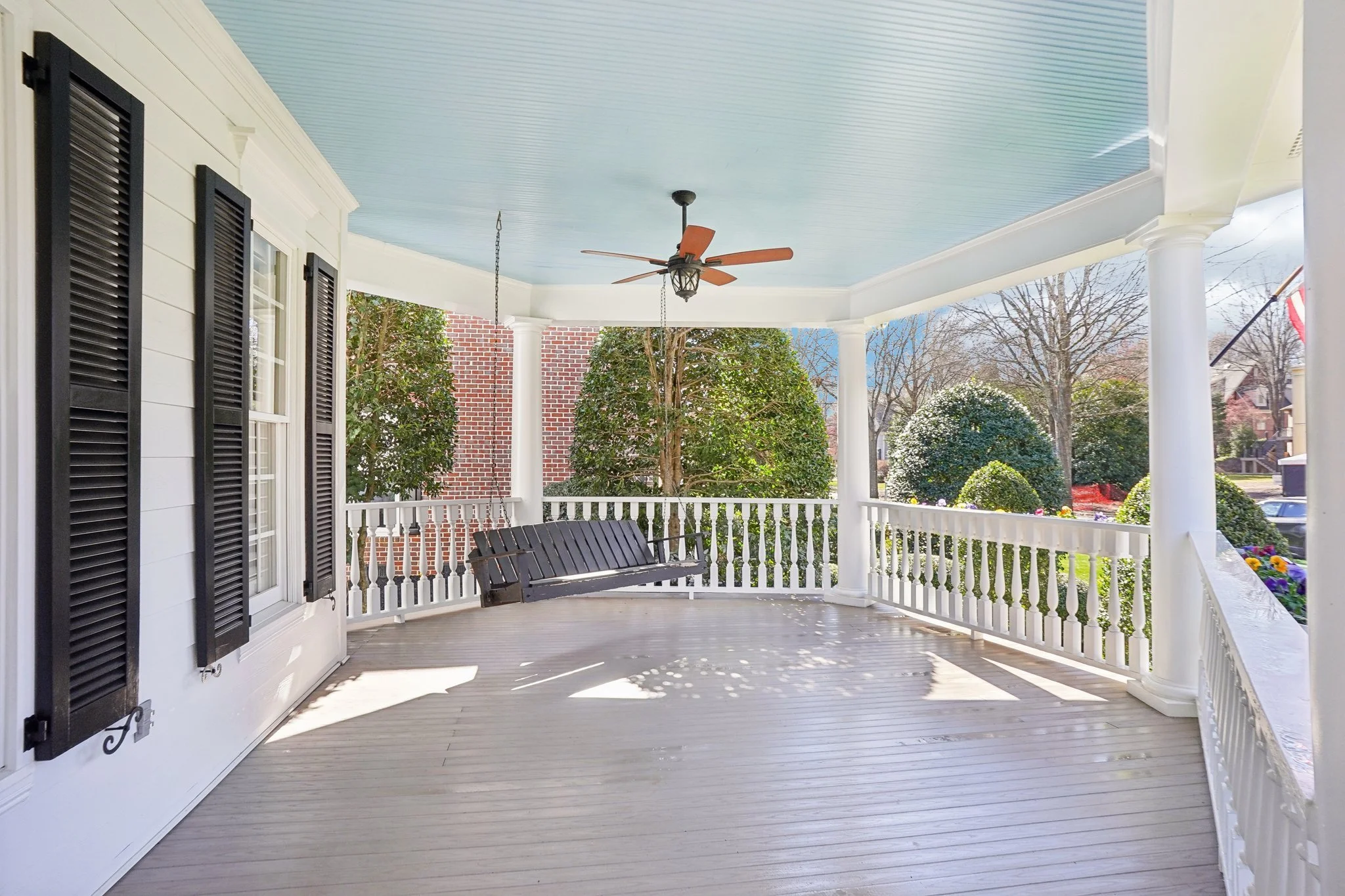 Empty front porch with white railings, black window shutters, and a black porch swing, surrounded by trees and bushes with a blue ceiling, ceiling fan, and sunlight casting shadows.