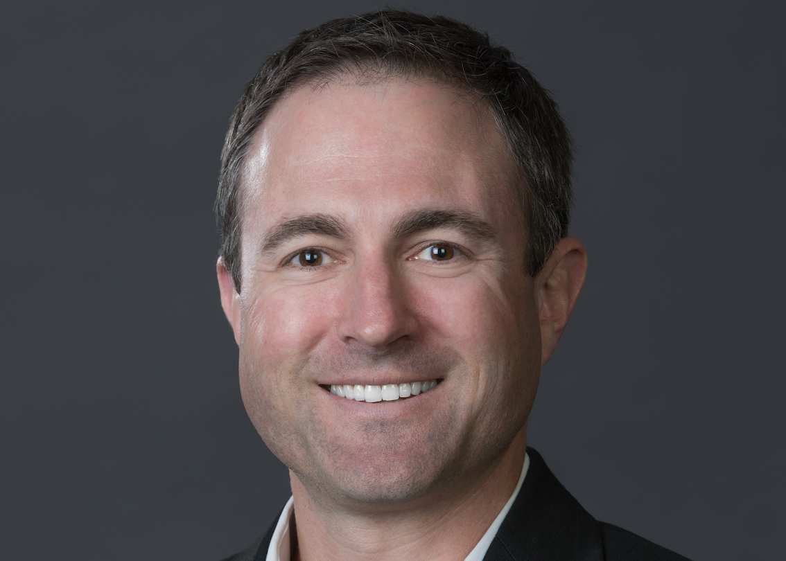 Portrait of a smiling man with short brown hair, dressed in a dark suit, white shirt, against a gray background.