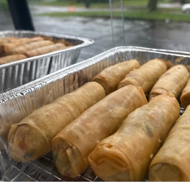 Rows of freshly fried egg rolls in aluminum trays at an outdoor food vendor stand, with a rainy street and trees in the background.
