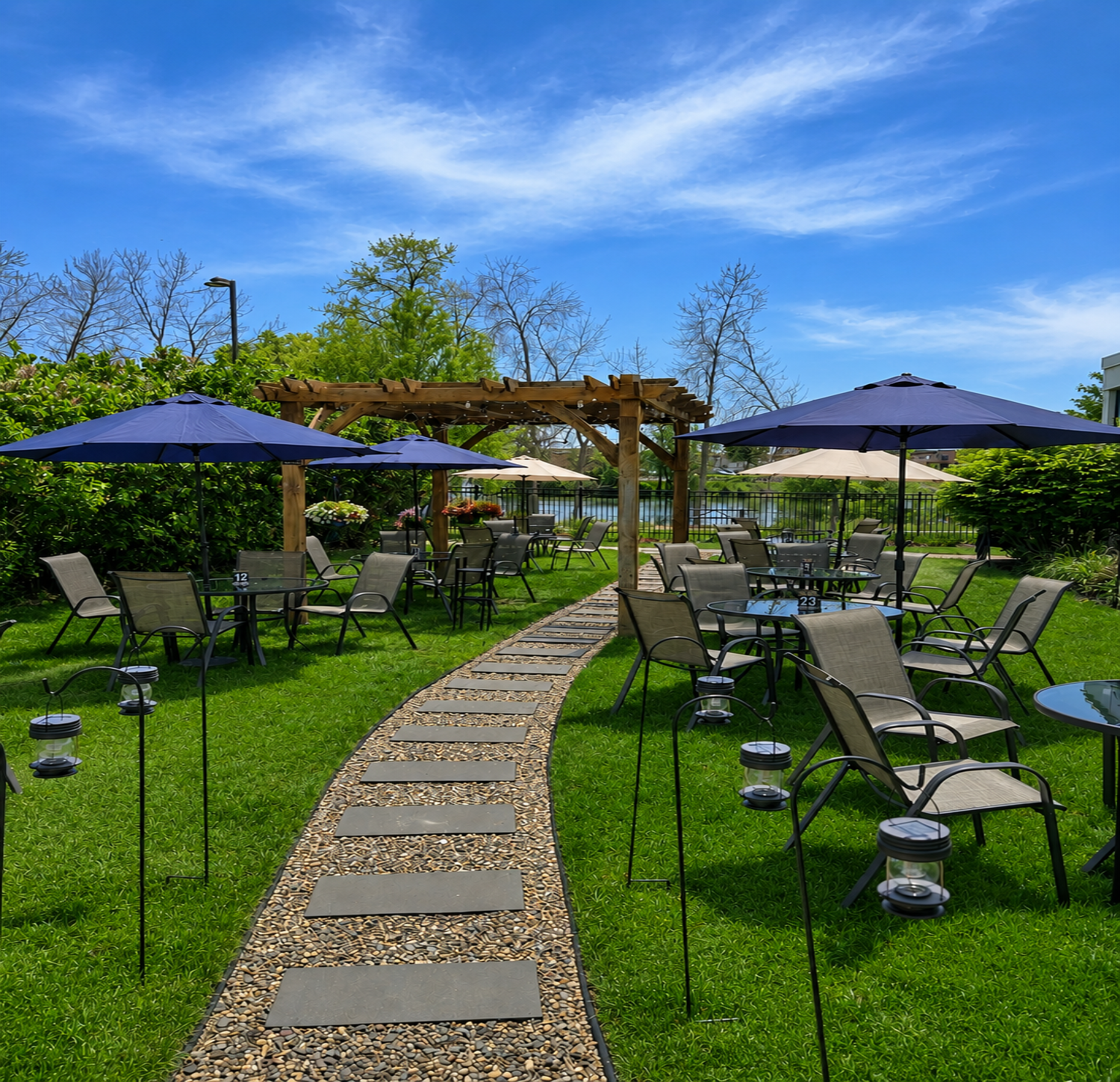 An outdoor patio area with multiple tables, chairs, and umbrellas on a green lawn, featuring a wooden pergola, a pathway with stepping stones, and a lake or pond in the background under a blue sky with some clouds.