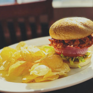 A plate with a sandwich and potato chips. The sandwich has a bun, fried chicken, lettuce, tomato, and onion.