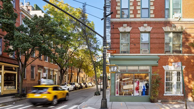 Vista di una strada urbana con edifici in mattoni rossi, un negozio con vetrina e un Taxi giallo che passa.
