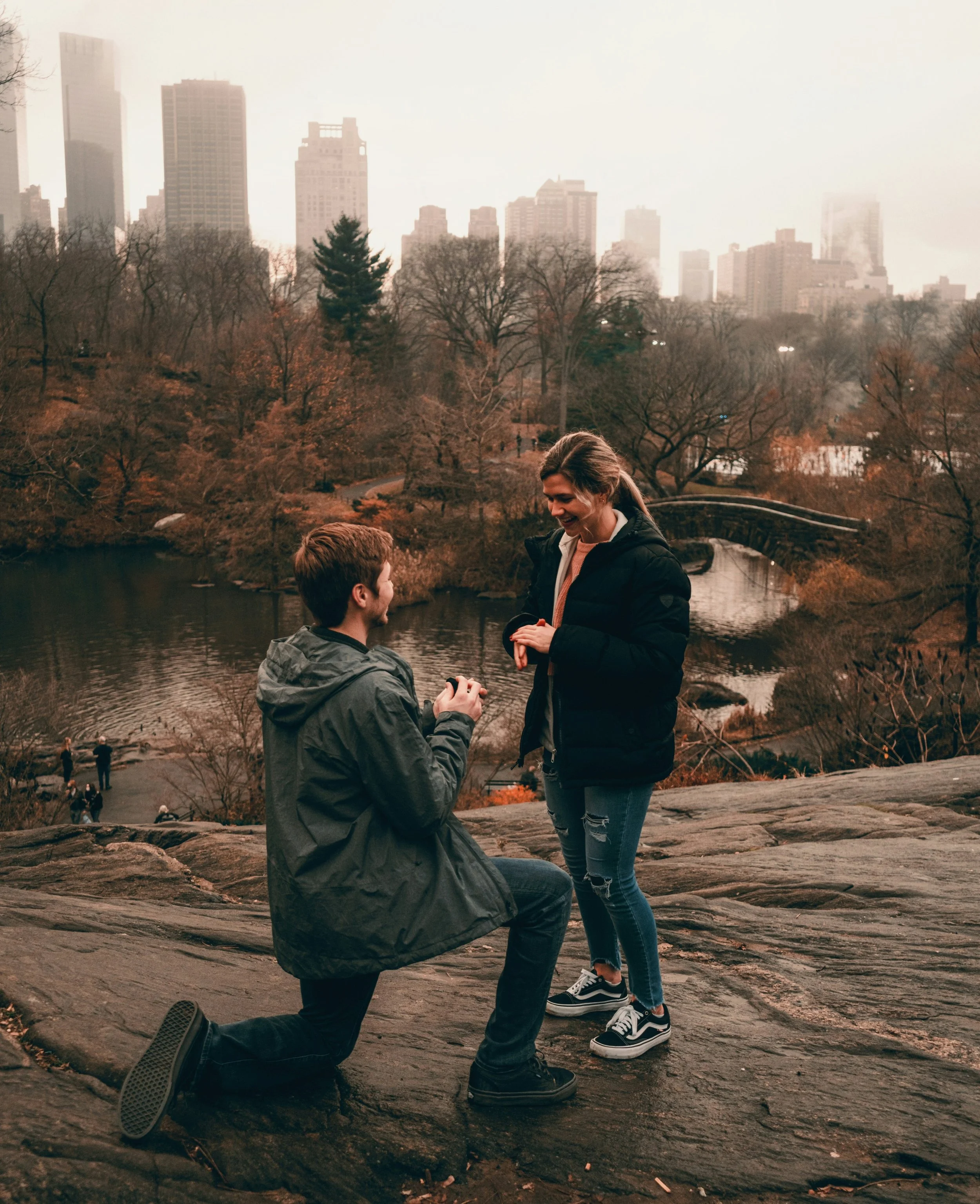 Un uomo chiede la mano di una donna in un parco con uno skyline urbano sullo sfondo.