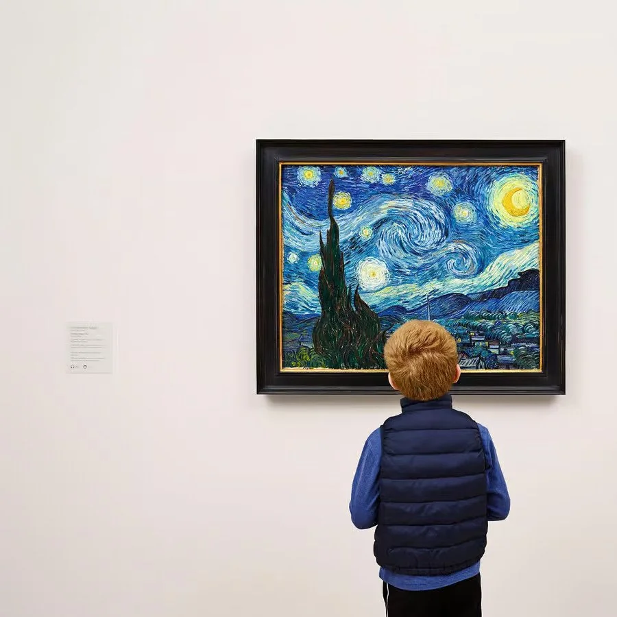 Un bambino che guarda il dipinto "La notte stellata" di Vincent van Gogh in un museo, con pareti bianche e un'etichetta descrittiva a sinistra.