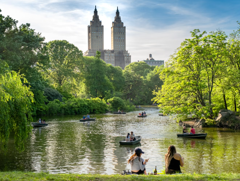 Due persone sedute sulle rive di un lago in un parco, mentre altre persone navigano in barche sullo stagno, con grattacieli visibili sullo sfondo.