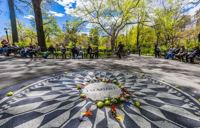 Cerchio di fiori e oggetti decorativi sull'Hot Pie di John Lennon a Central Park, New York, con persone sedute e camminamenti nel parco e alberi verdi sotto un cielo blu.