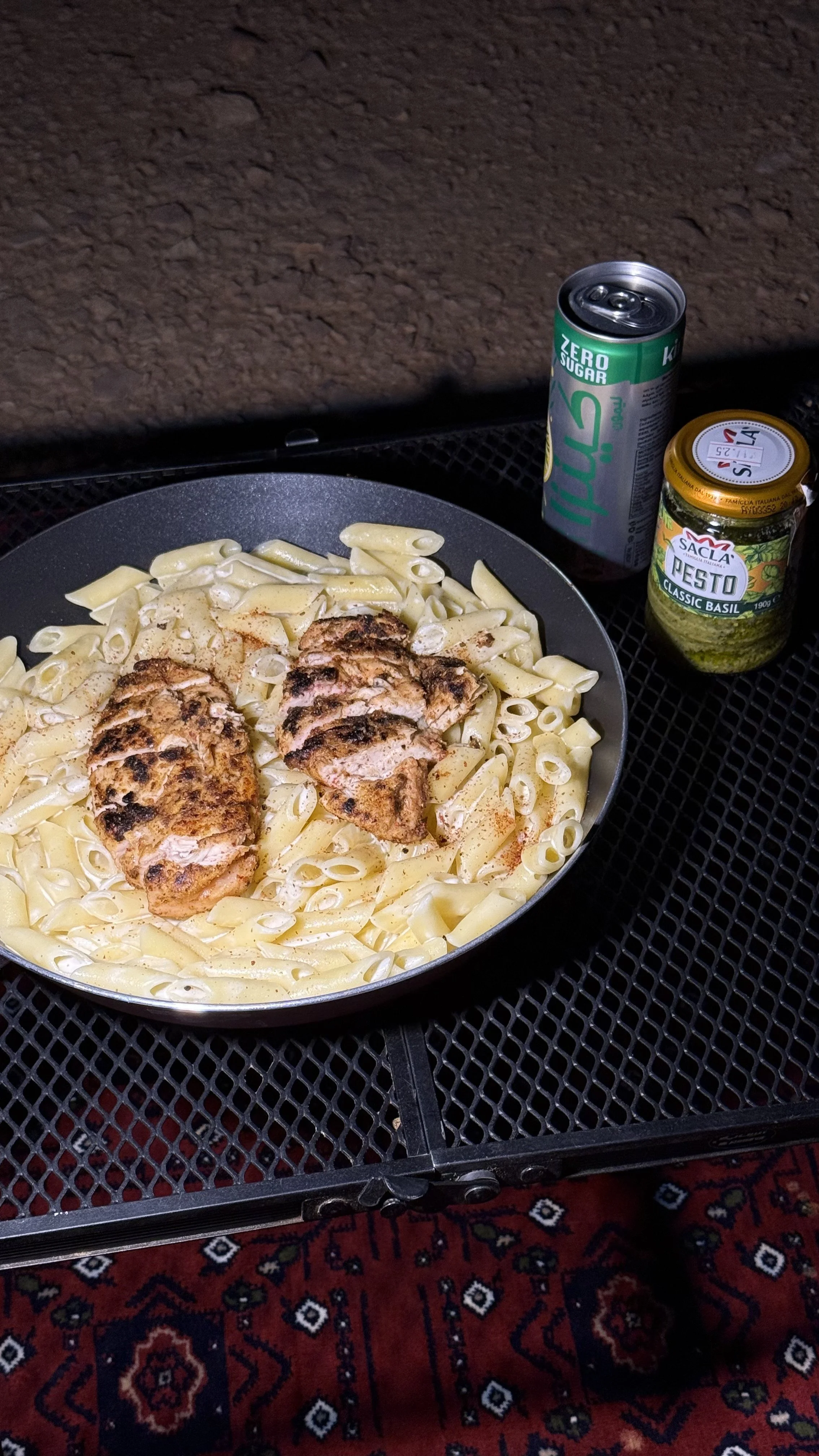 Cooked pasta with grilled chicken slices in a black skillet, accompanied by a can of zero sugar lemon soda and a jar of basil pesto, on a metal outdoor table with a patterned rug underneath.