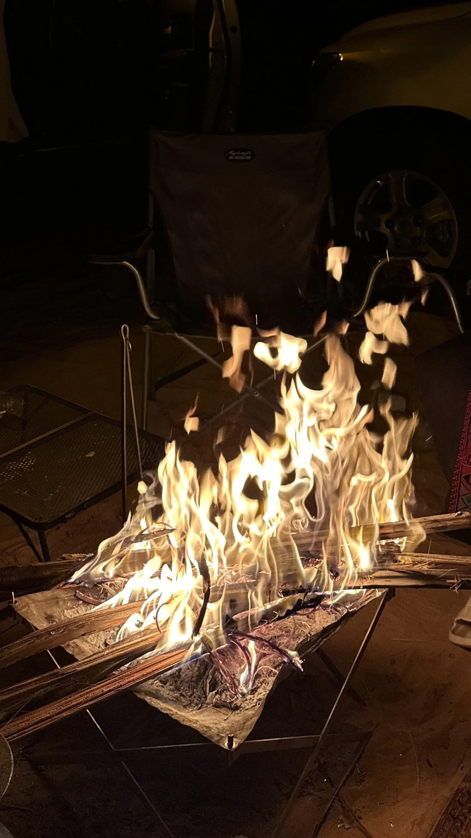 A campfire with flames burning on firewood sticks, with a black camping chair and parked cars visible in the dark background.