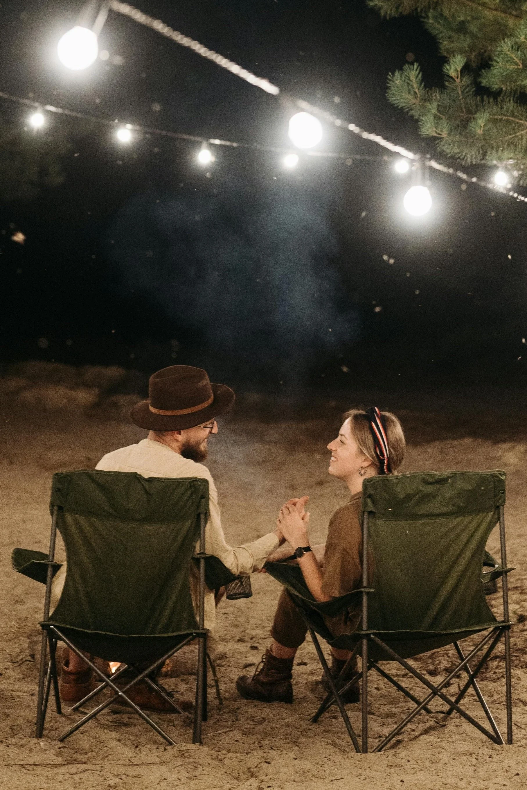 A man and woman sitting in outdoor camping chairs holding hands at night, with string lights above and trees in the background.