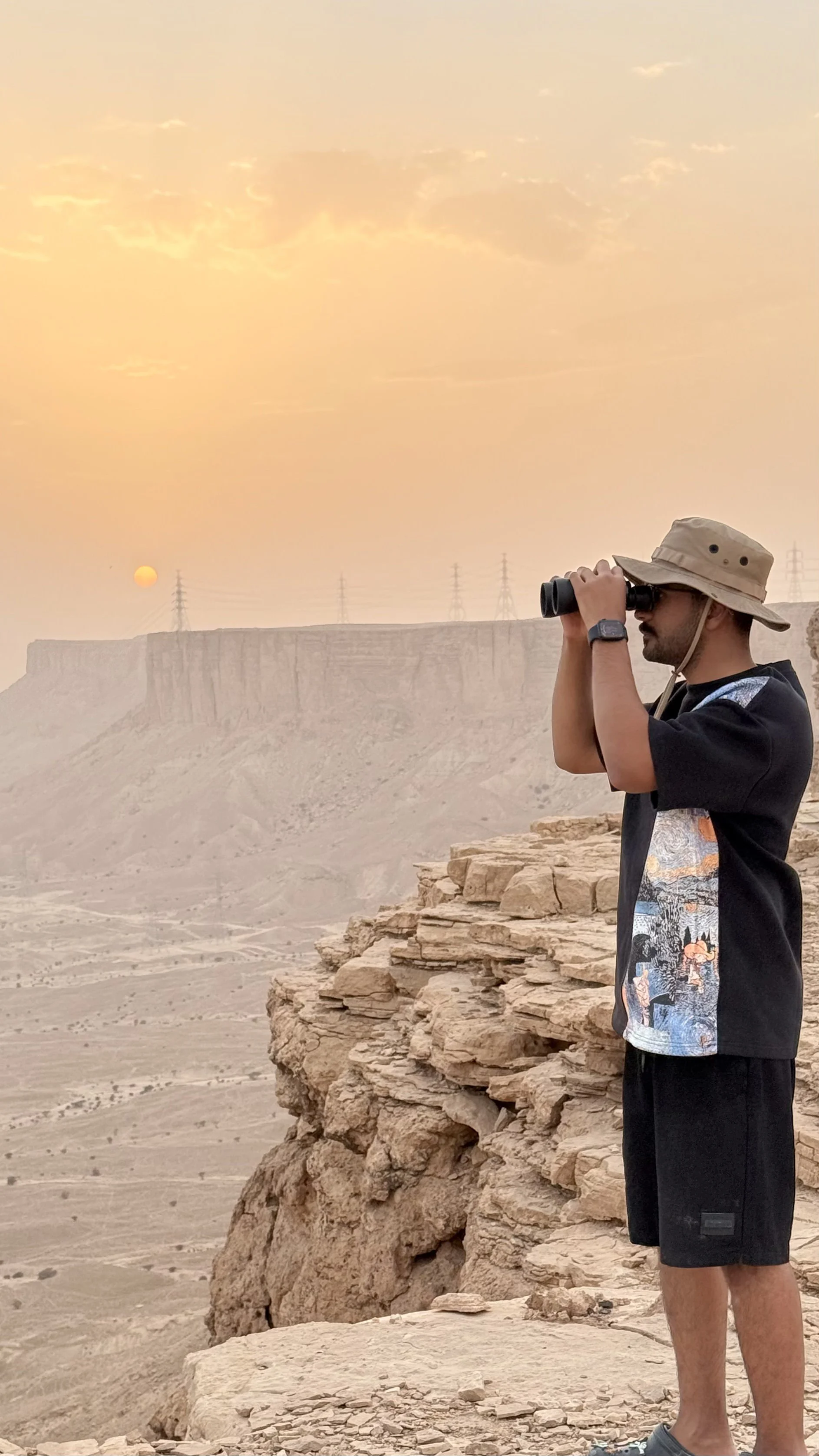 A man standing on rocky terrain in a desert landscape at sunset, looking through binoculars, wearing a sun hat, black t-shirt, and shorts.
