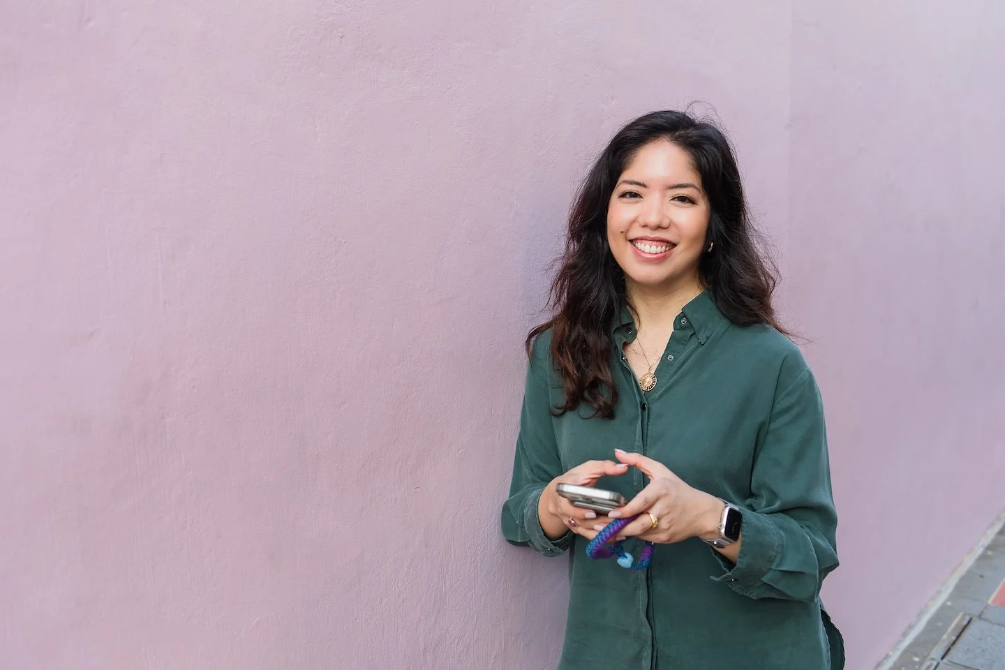 A smiling woman with long, wavy dark hair, wearing a green button-up shirt, holding a smartphone, standing against a pink wall.