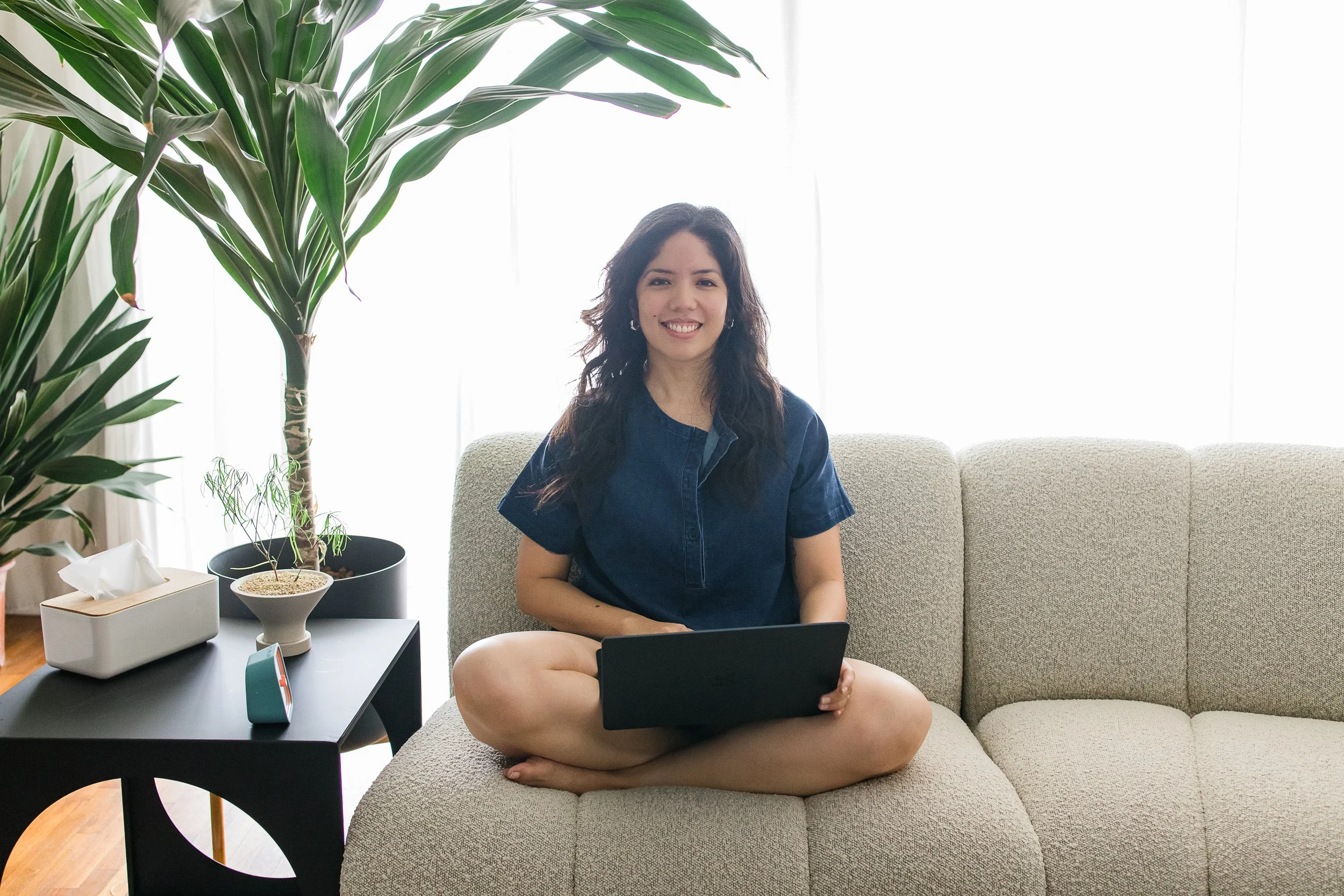 A young woman sitting cross-legged on a beige sofa, smiling, with a laptop on her lap, in a bright living room with a large houseplant and a side table with a small plant, tissue box, and speaker.