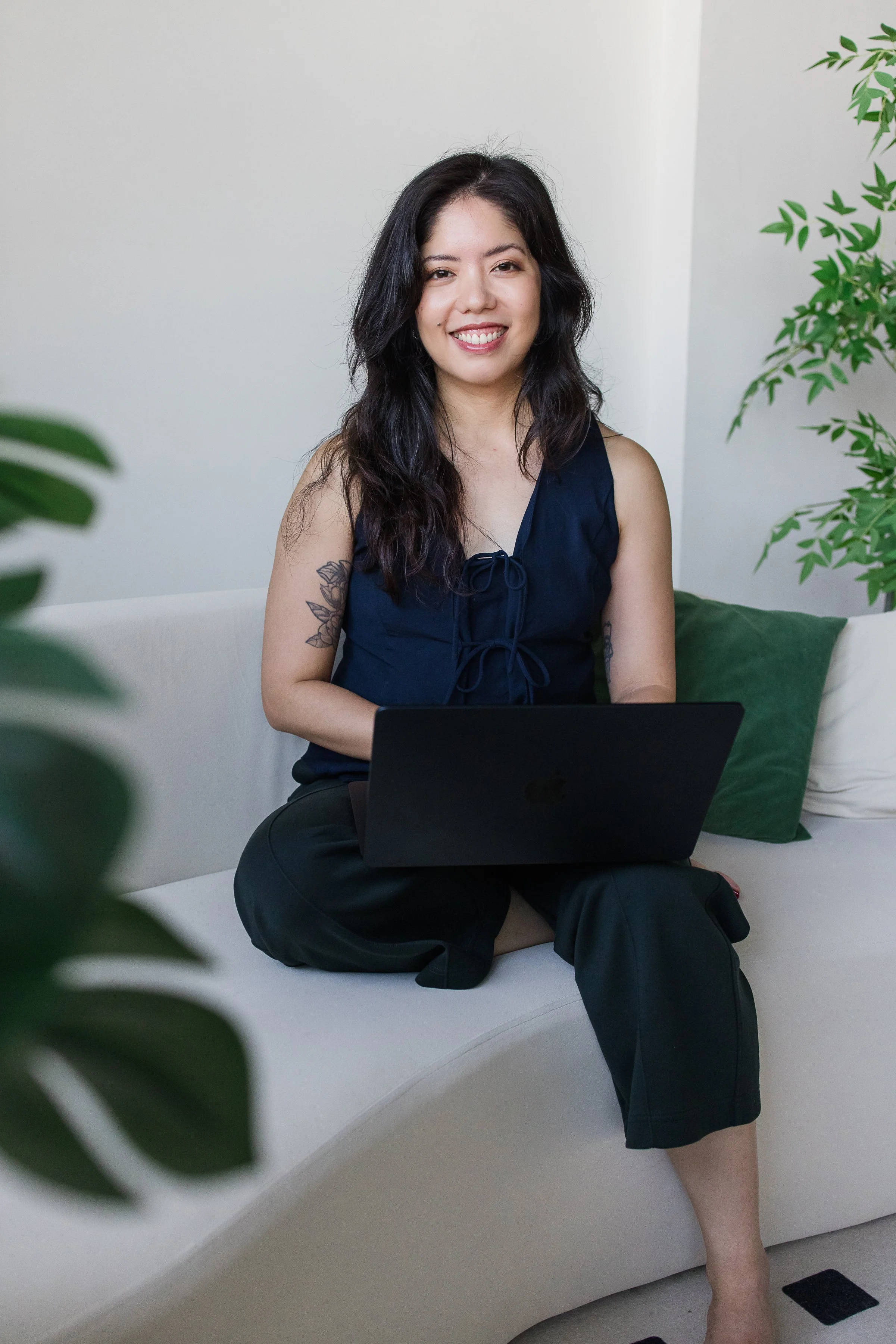 A woman sitting on a white sofa with a laptop, smiling, surrounded by green plants.