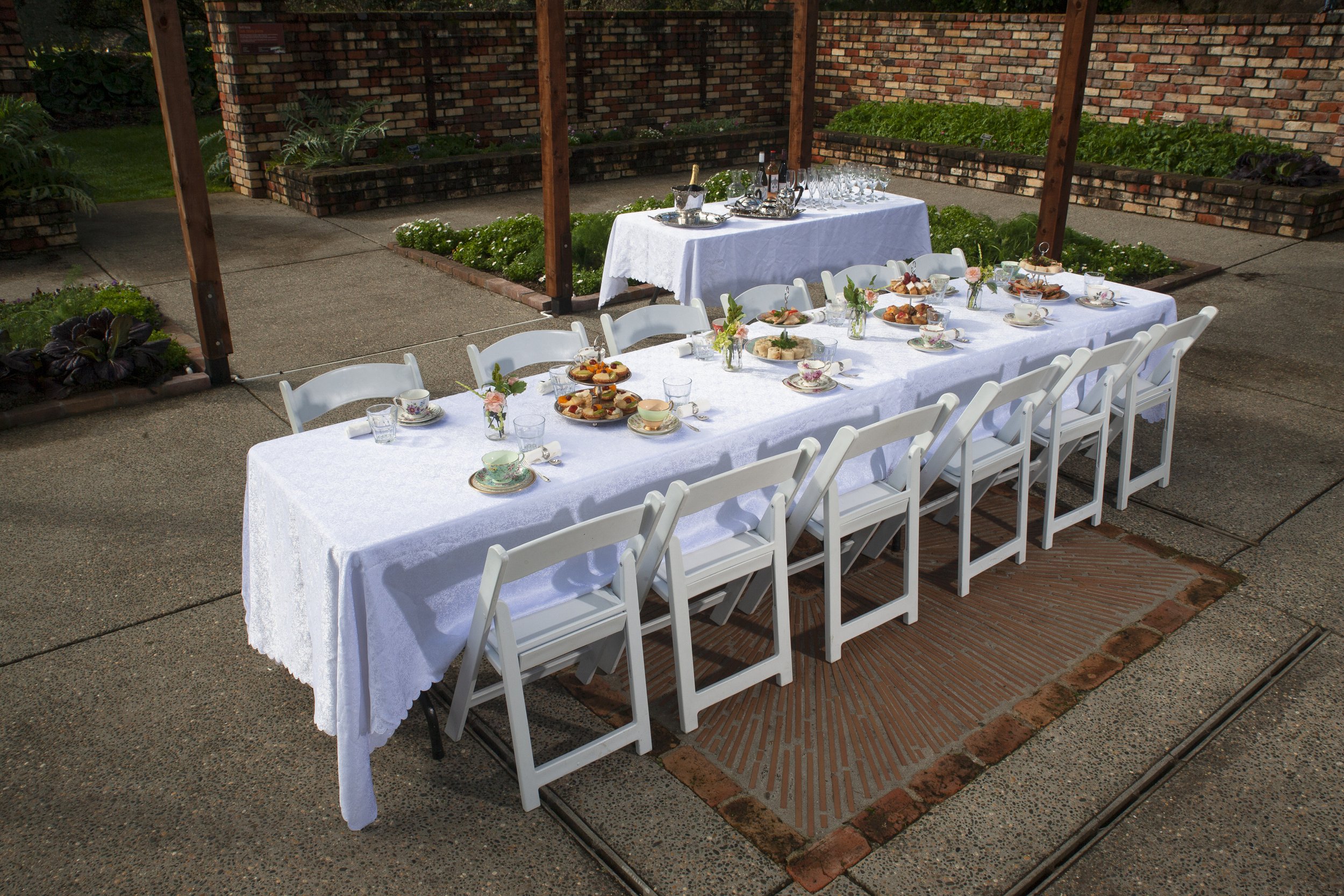 Outdoor dining setup with a long table covered with a white tablecloth, set with teacups, plates of pastries, and floral centerpieces, surrounded by white folding chairs, and a secondary table with glasses and beverages in the background.