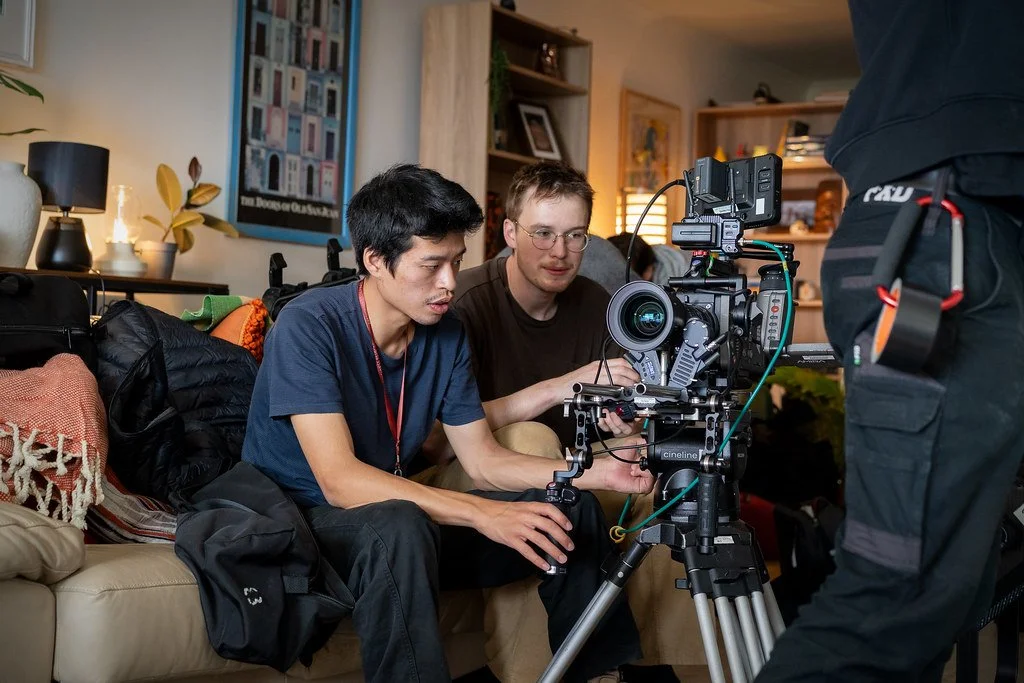 Two men operating a professional film camera on a tripod in a cozy living room with a sofa, bookshelves, and wall art.