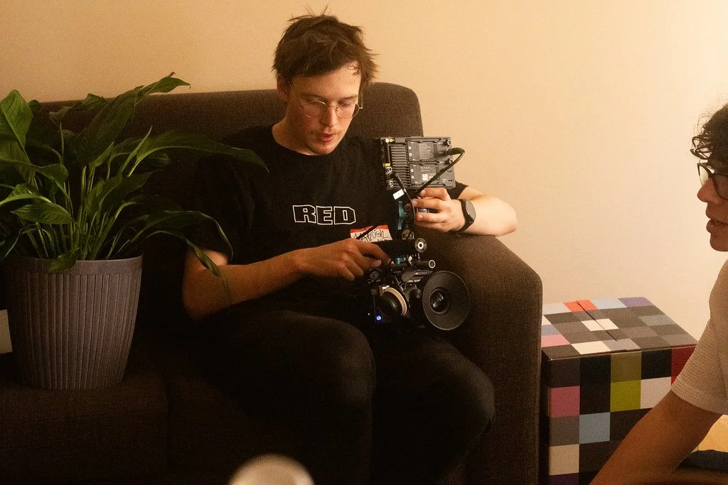 A young man with glasses sitting on a brown couch, holding a camera rig. There is a potted plant with large green leaves on his left and a colorful checkered box on his right, in a room with beige walls.