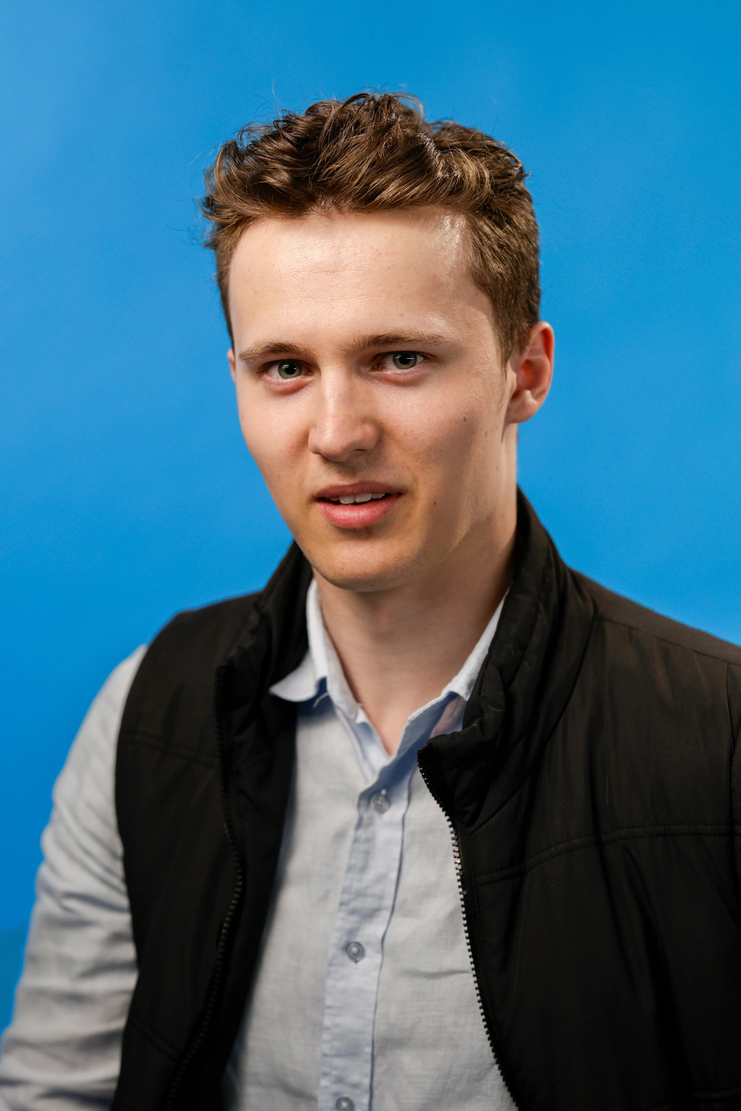 A young man with light skin, short wavy brown hair, and blue eyes, wearing a white collared shirt and a black vest, standing in front of a blue background.