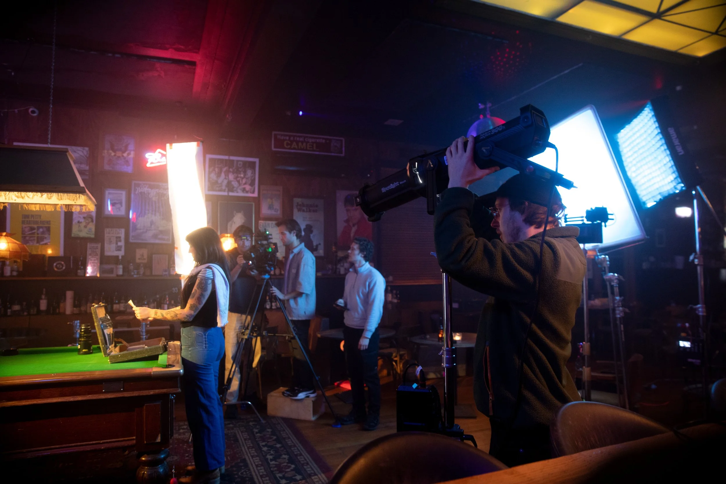 People filming and taking photos in a dimly lit bar or restaurant with neon signs, paintings, and a pool table in the foreground.