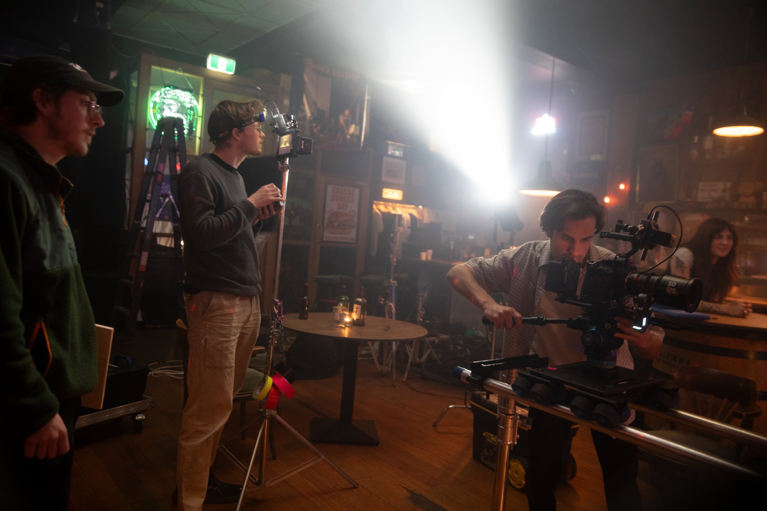 Film crew setting up camera equipment inside a dimly lit restaurant or bar with neon signs and people in the background.