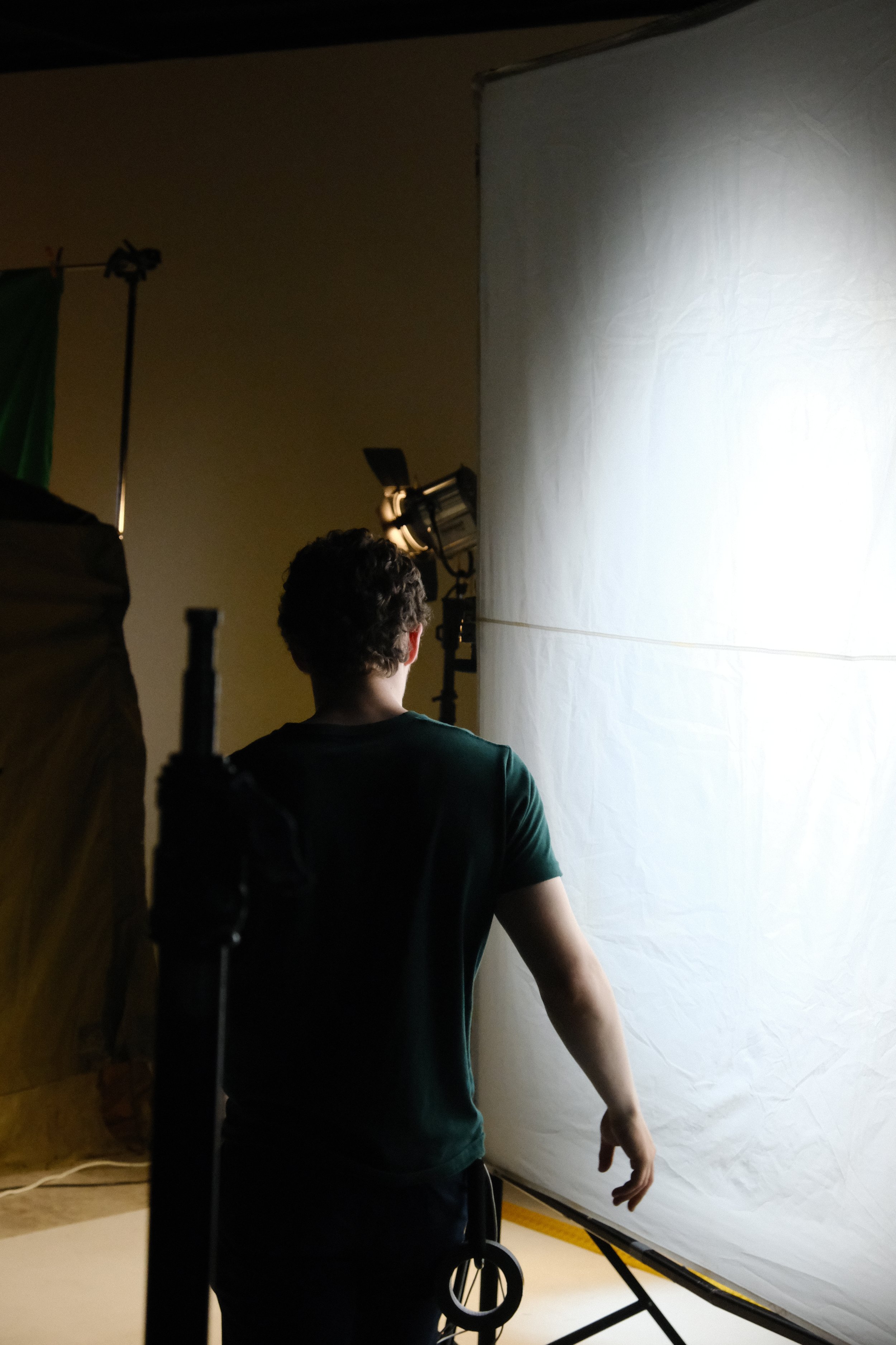 A person stands with their back to the camera in front of a white sheet or backdrop, with professional studio lighting aimed at them.