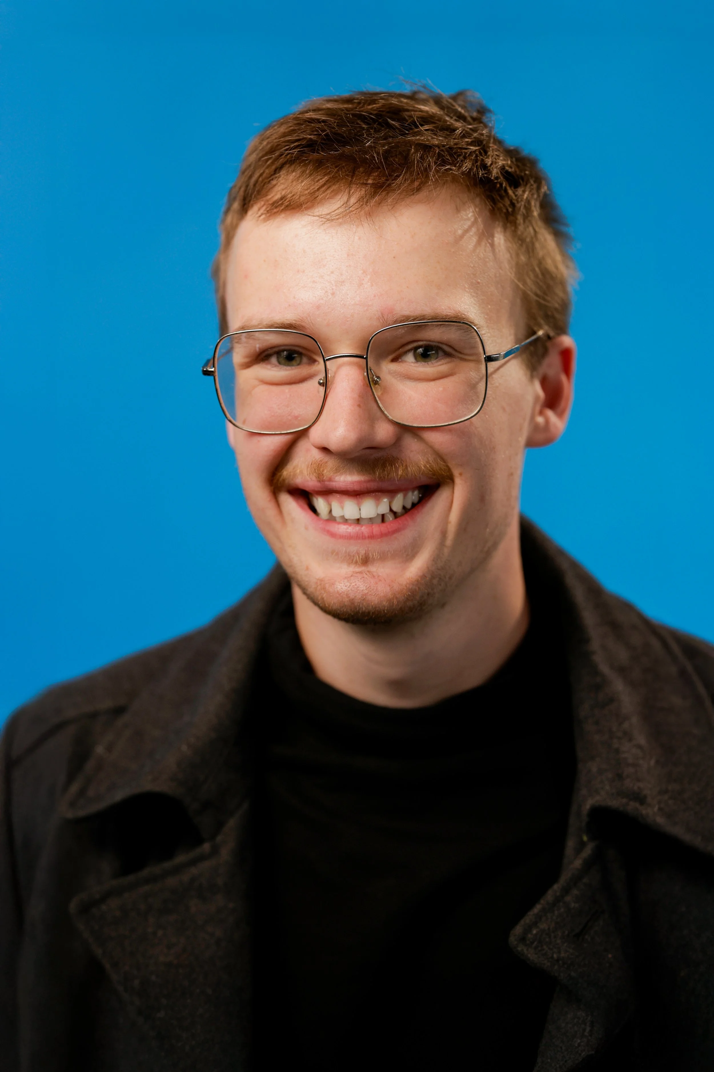 A young man with short brown hair, glasses, a light beard, and a big smile, wearing a black turtleneck and dark jacket, against a blue background.