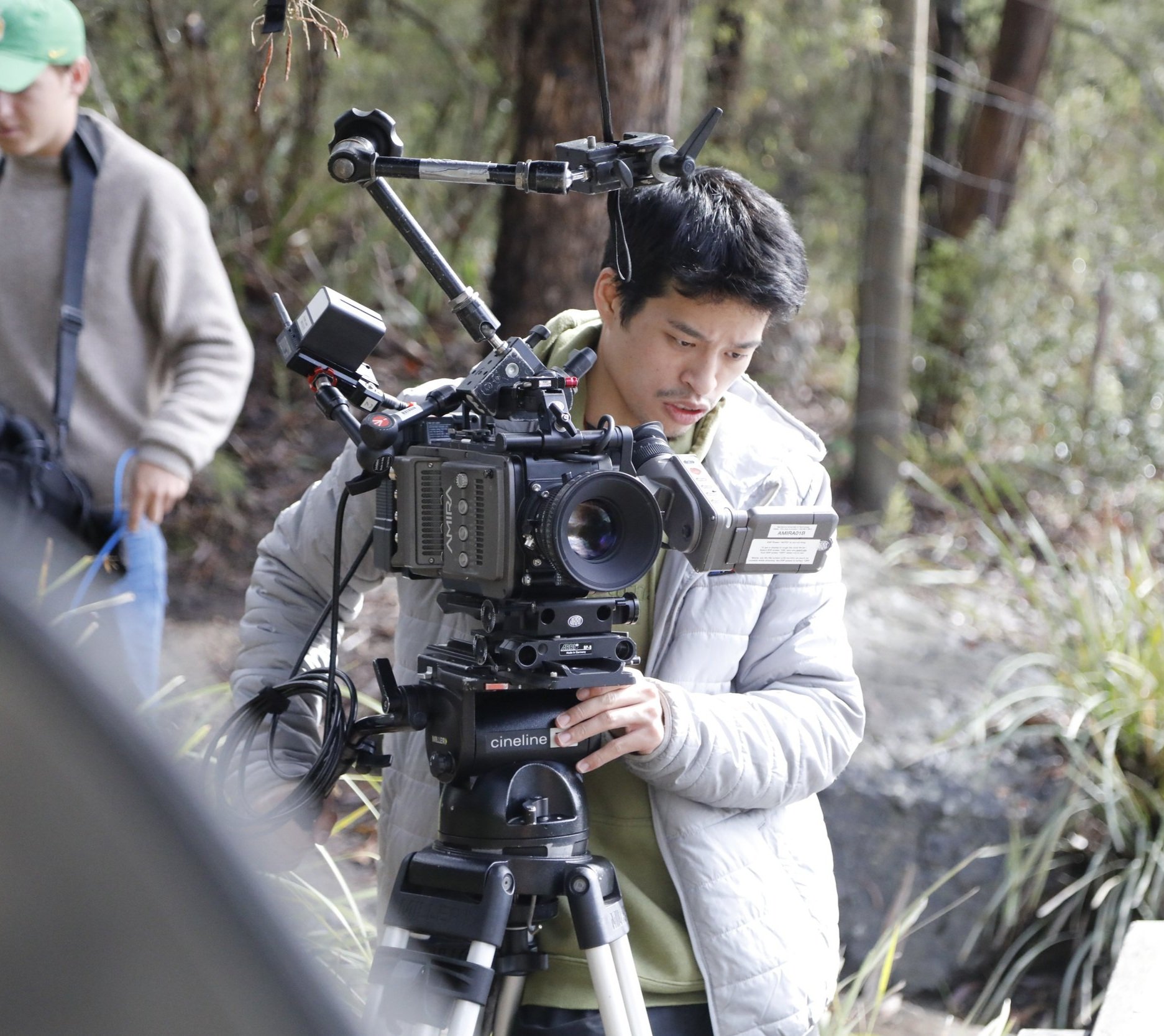 A person operating a professional video camera on a tripod outdoors in a wooded area, with another person partially visible on the left side of the image.