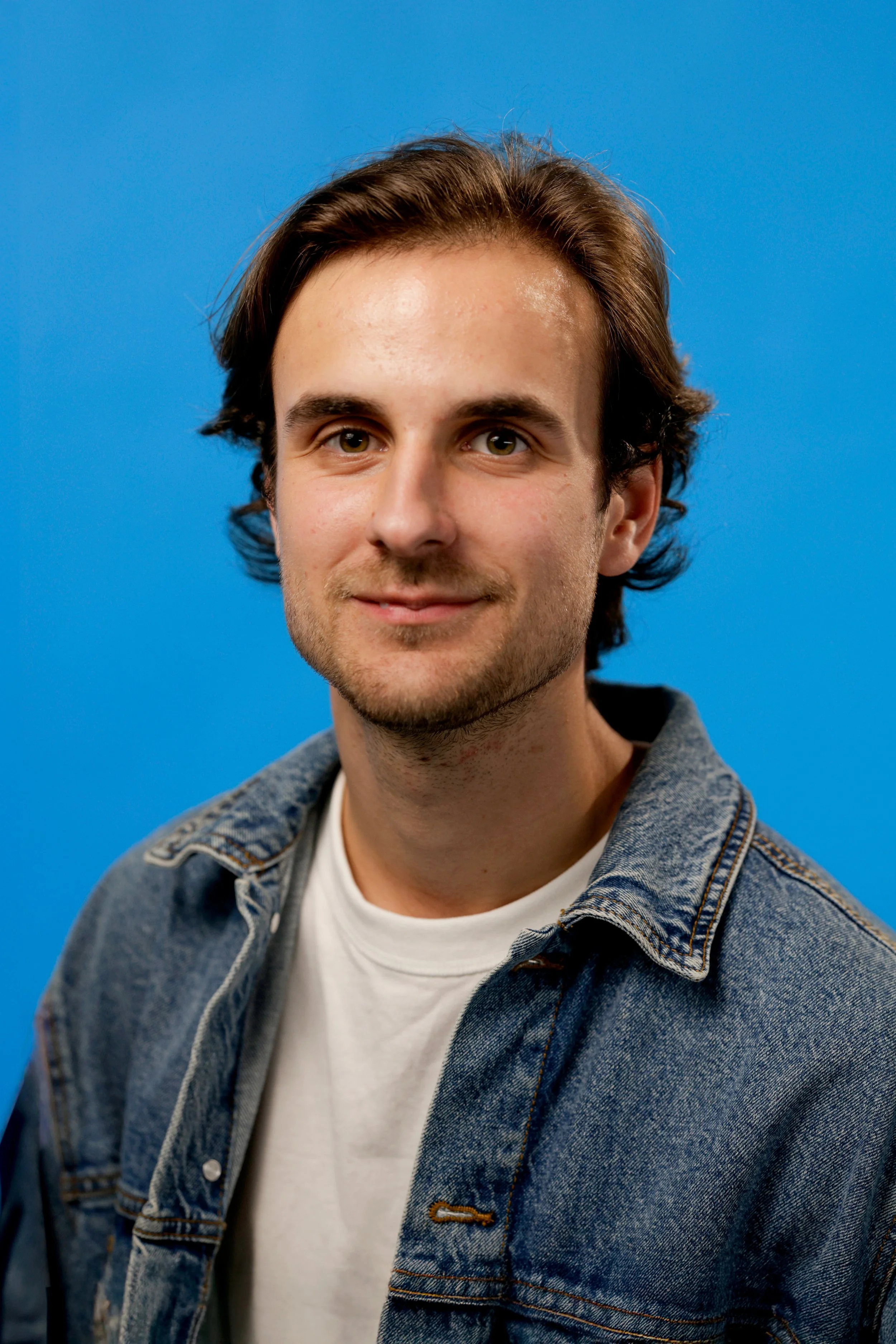 A young man with brown hair and a slight beard, wearing a white t-shirt and denim jacket, standing in front of a blue background.