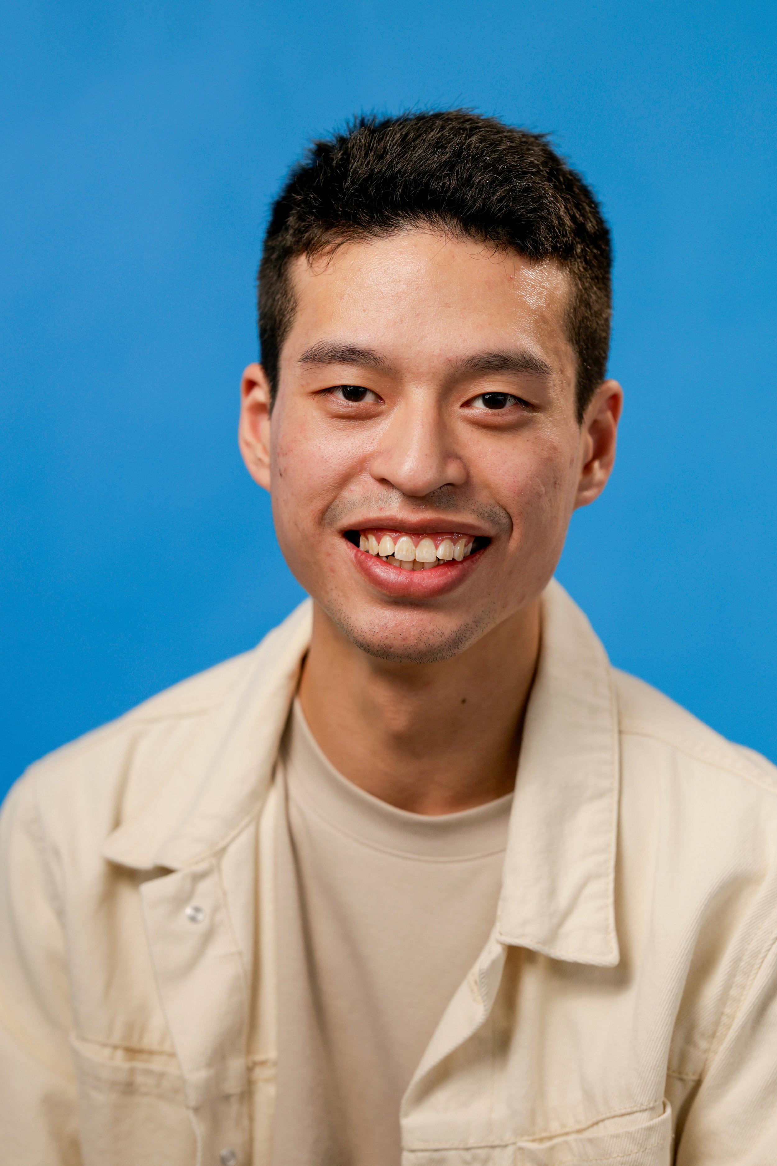 A young man with short dark hair, smiling, wearing a beige shirt against a blue background.