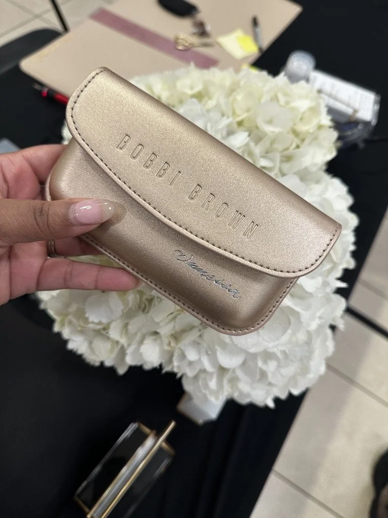 A person holding a metallic gold Bobbi Brown makeup pouch with the brand name embossed on it, in front of a bouquet of white flowers on a black table.
