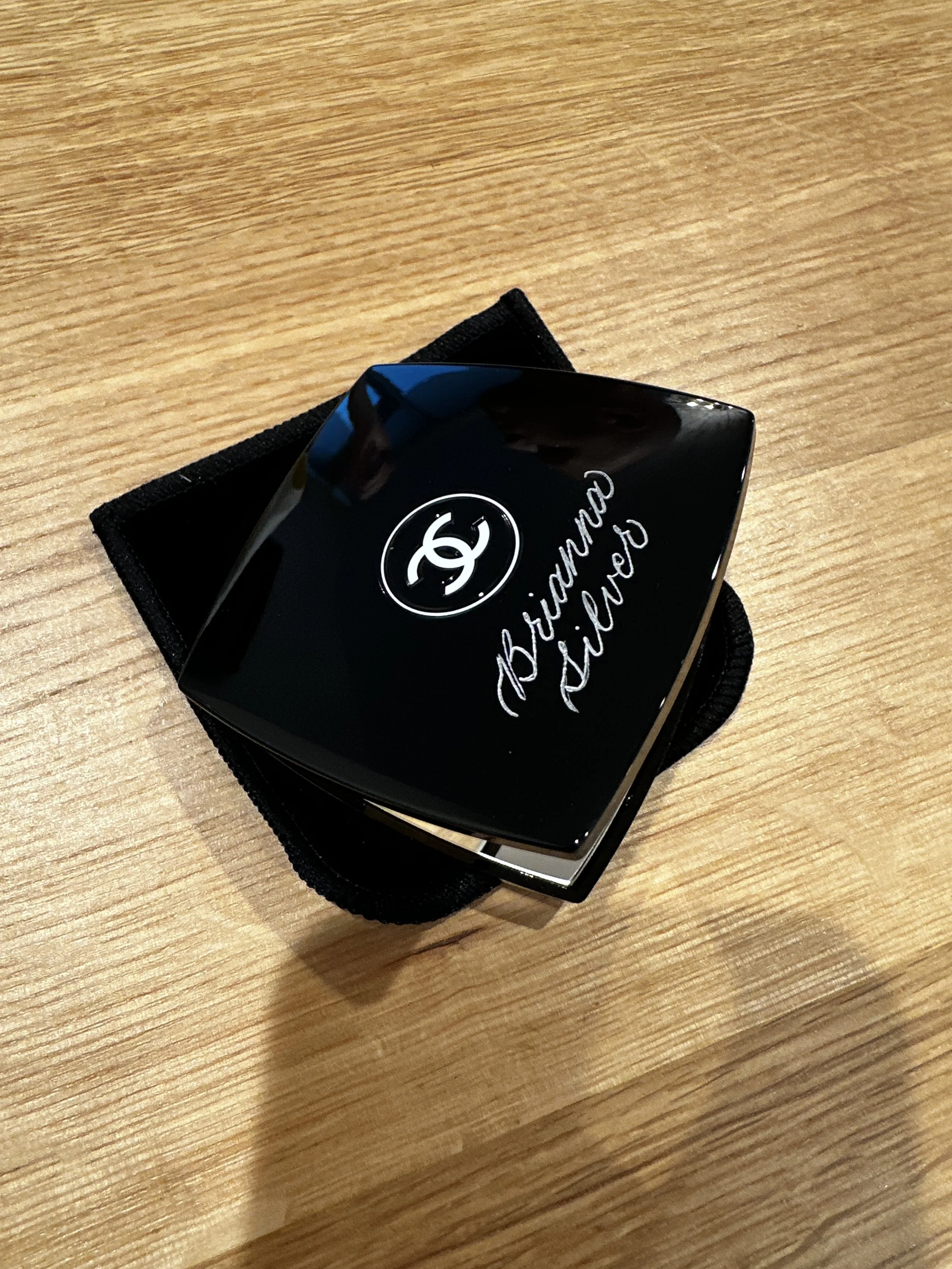A shiny black Chanel compact mirror with the Chanel logo and the words 'Brianna Silver' written in calligraphy on the top, resting on a black cloth on a wooden surface.
